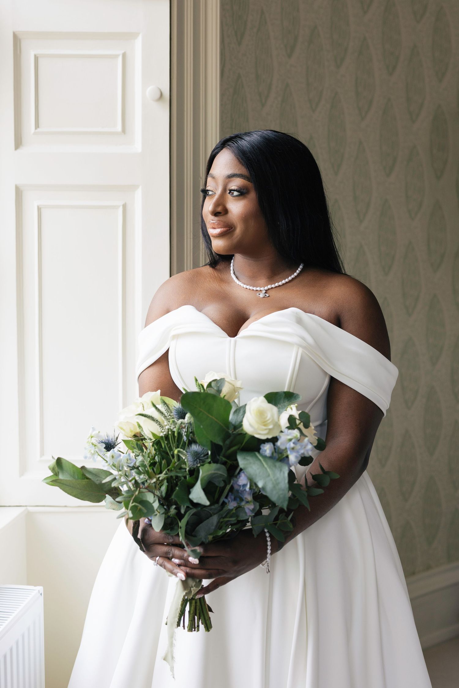 An elegant bride in an off-shoulder white gown holding a bouquet of white and blue flowers near a bright window Blair Castle Wedding Bride