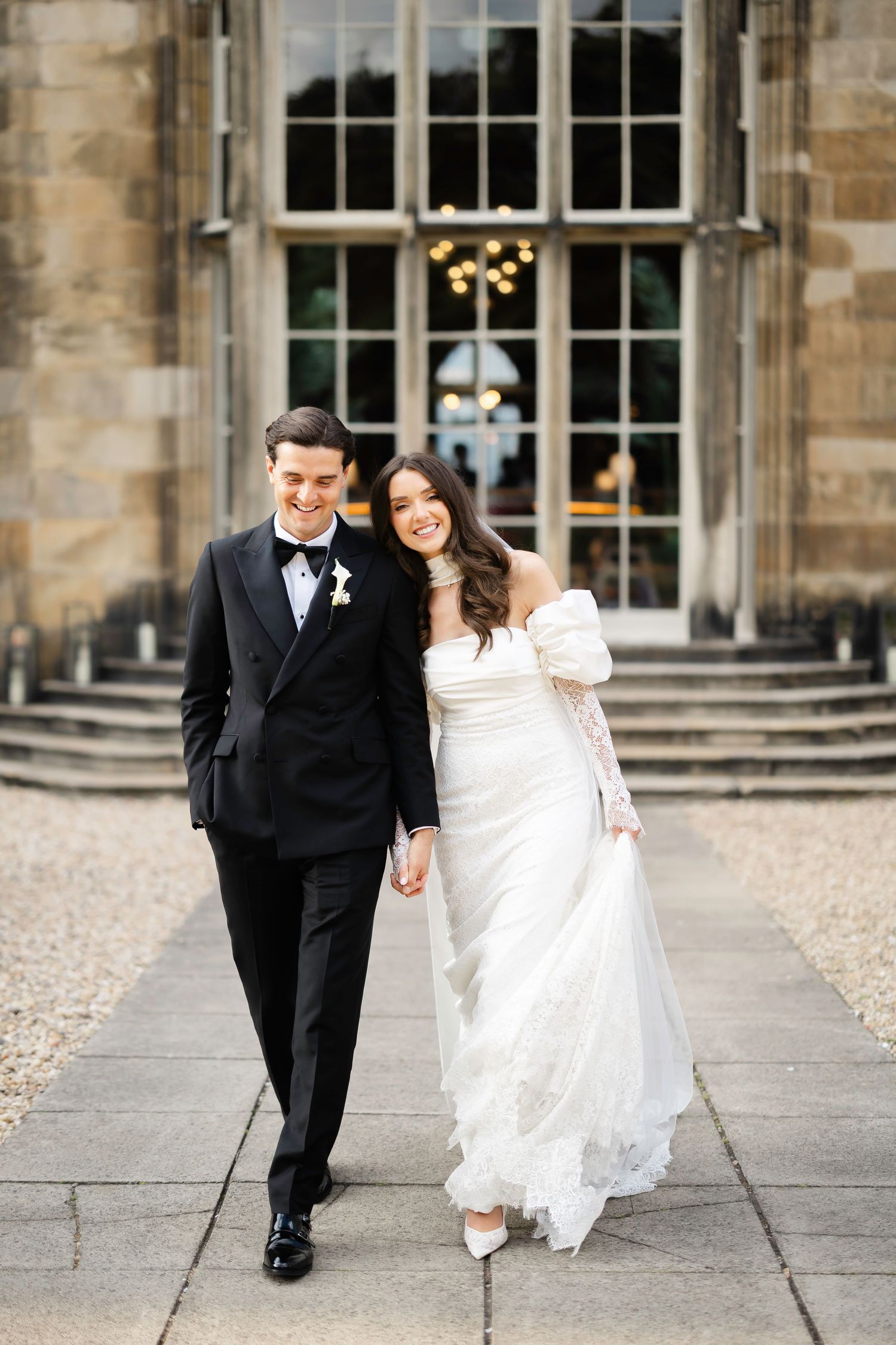 A bride and groom walk hand in hand outside a grand stone venue, she in a white off-shoulder gown, he in a black tuxedo.