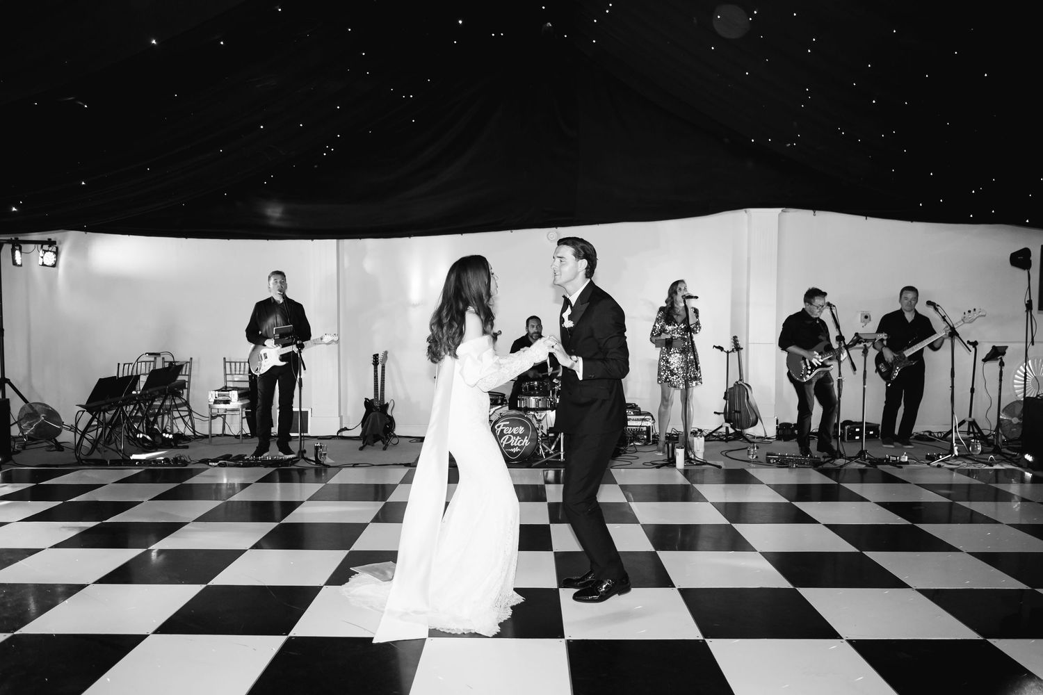 Couple shares first dance on checkered floor with live band performing behind them at elegant wedding reception.