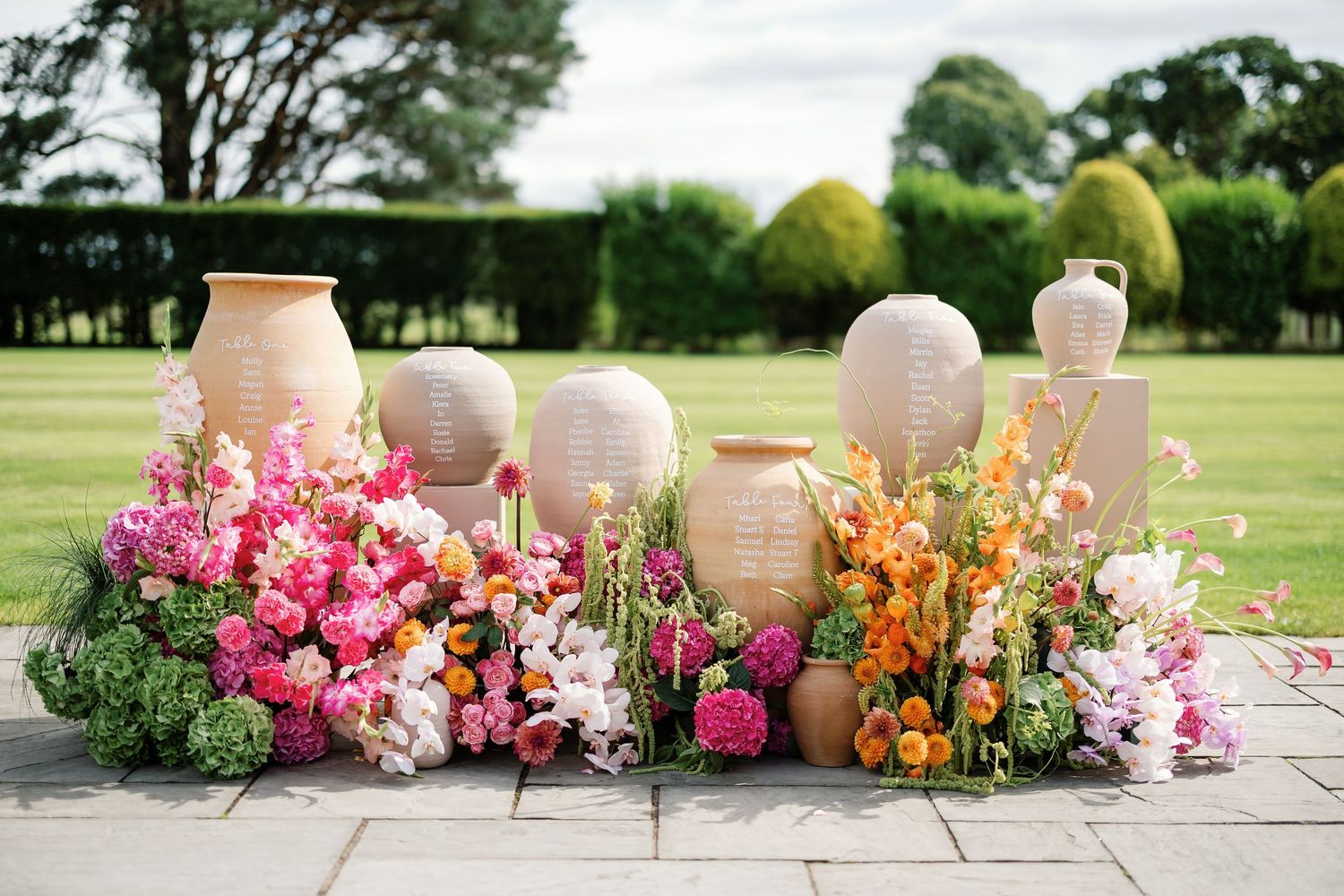 Panoramic view of colorful floral arrangements with ceramic vases displayed outdoors on a stone patio against a green lawn.