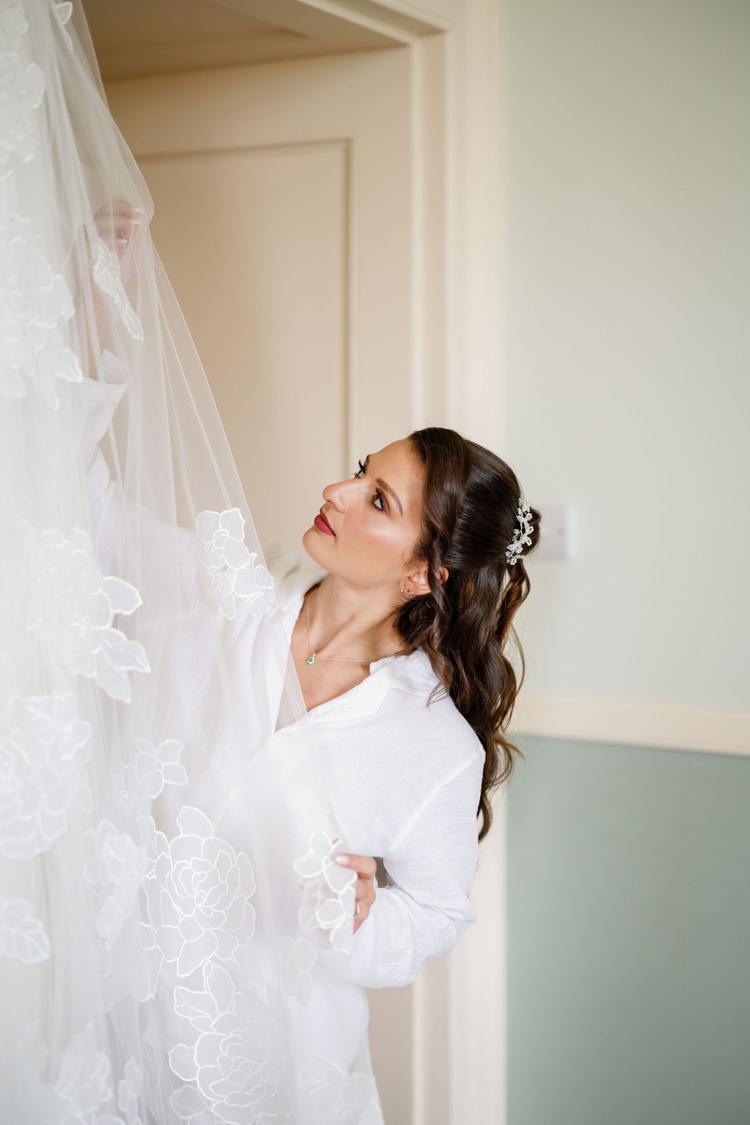 A bride in a white dress looks up joyfully while standing near delicate lace curtains in a soft-lit room.