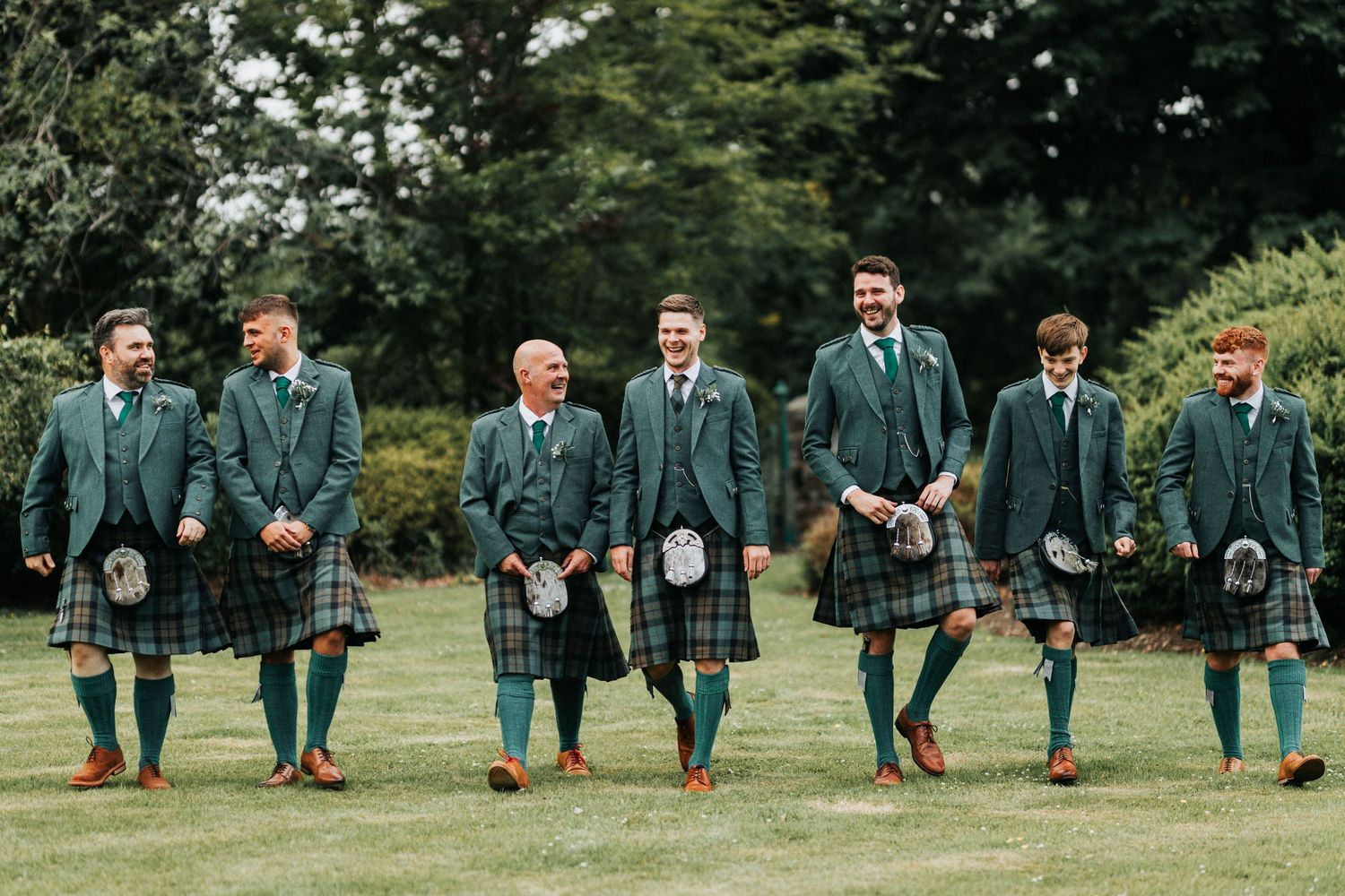 Wedding party in traditional Scottish green kilts and jackets stand in a line on grass with trees in background.