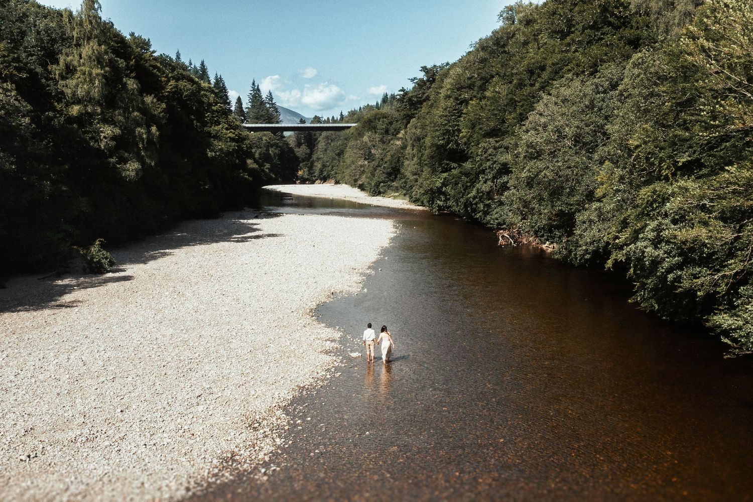 Tranquil river scene with clear waters flowing beneath a bridge between forested riverbanks on a sunny day.