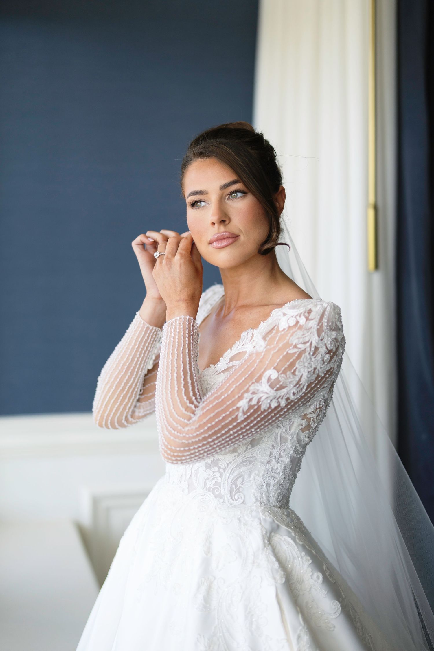 Elegant bride in long sleeve lace wedding gown with pearl beading poses near window in romantic bridal portrait. Trump Turnberry wedding