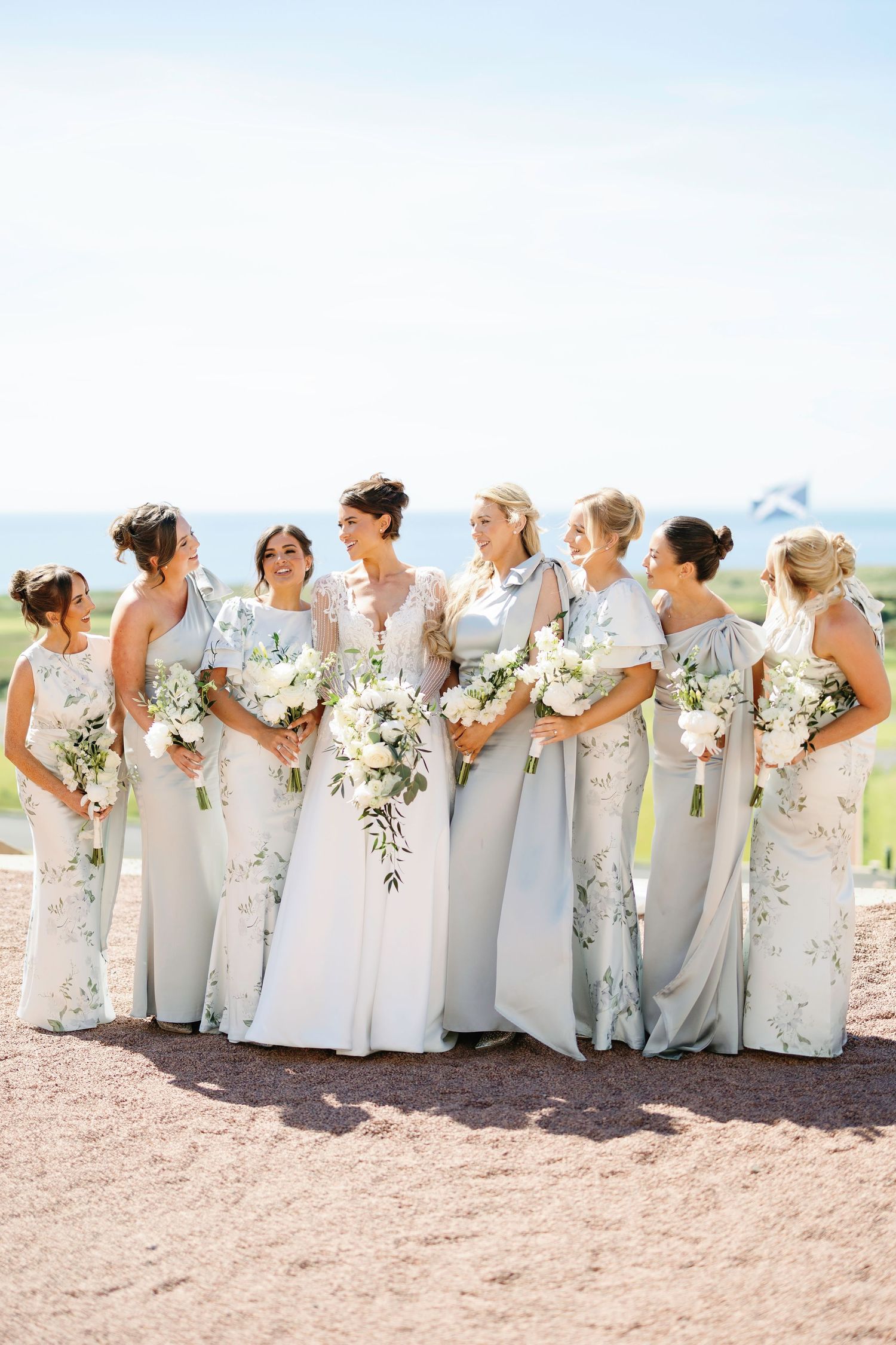 Bridesmaids in soft floral dresses stand together holding white bouquets against coastal backdrop at Trump Turnberry