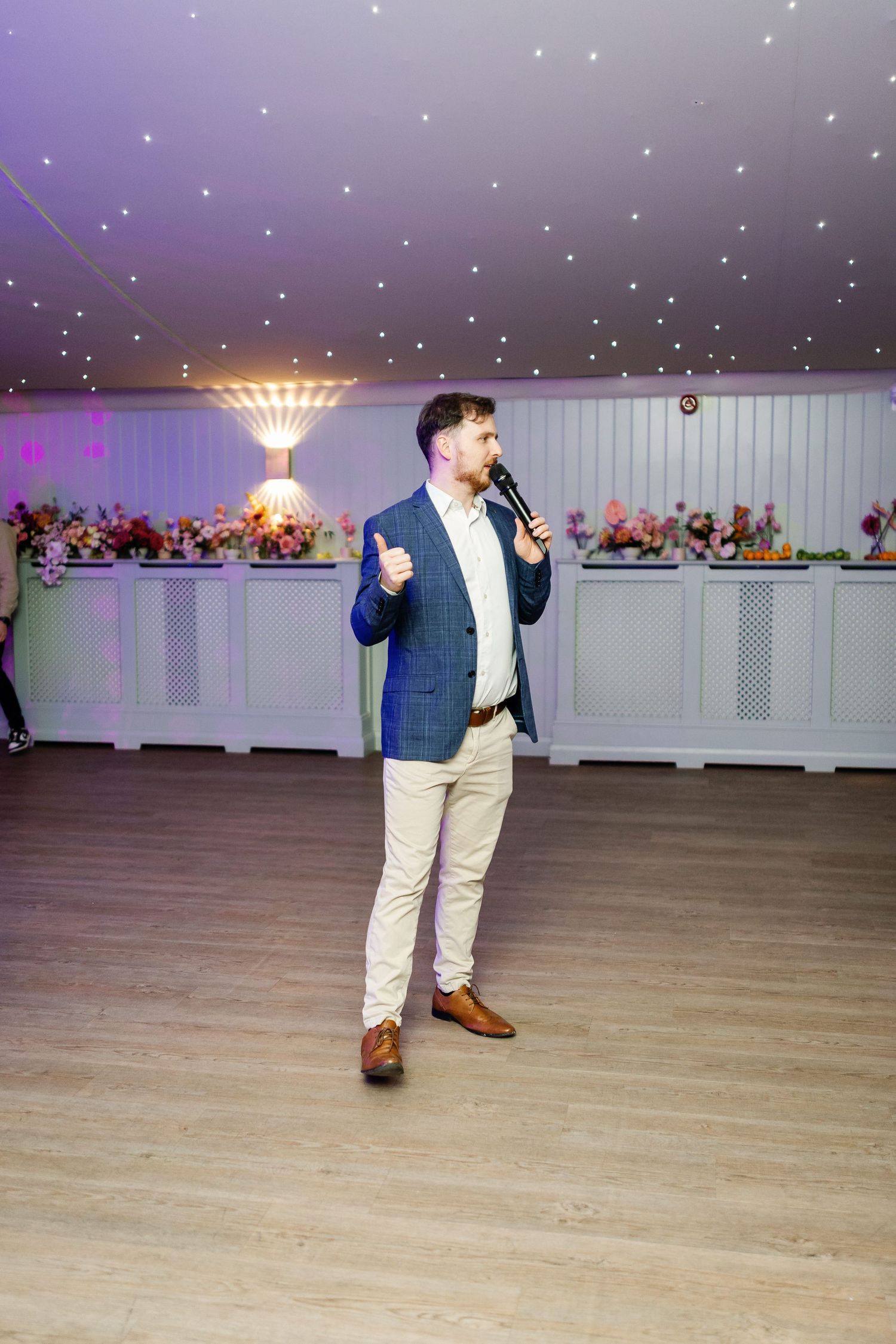 A person in a navy blazer and khaki pants performs a dance move on a wooden floor at an event venue with purple lighting.
