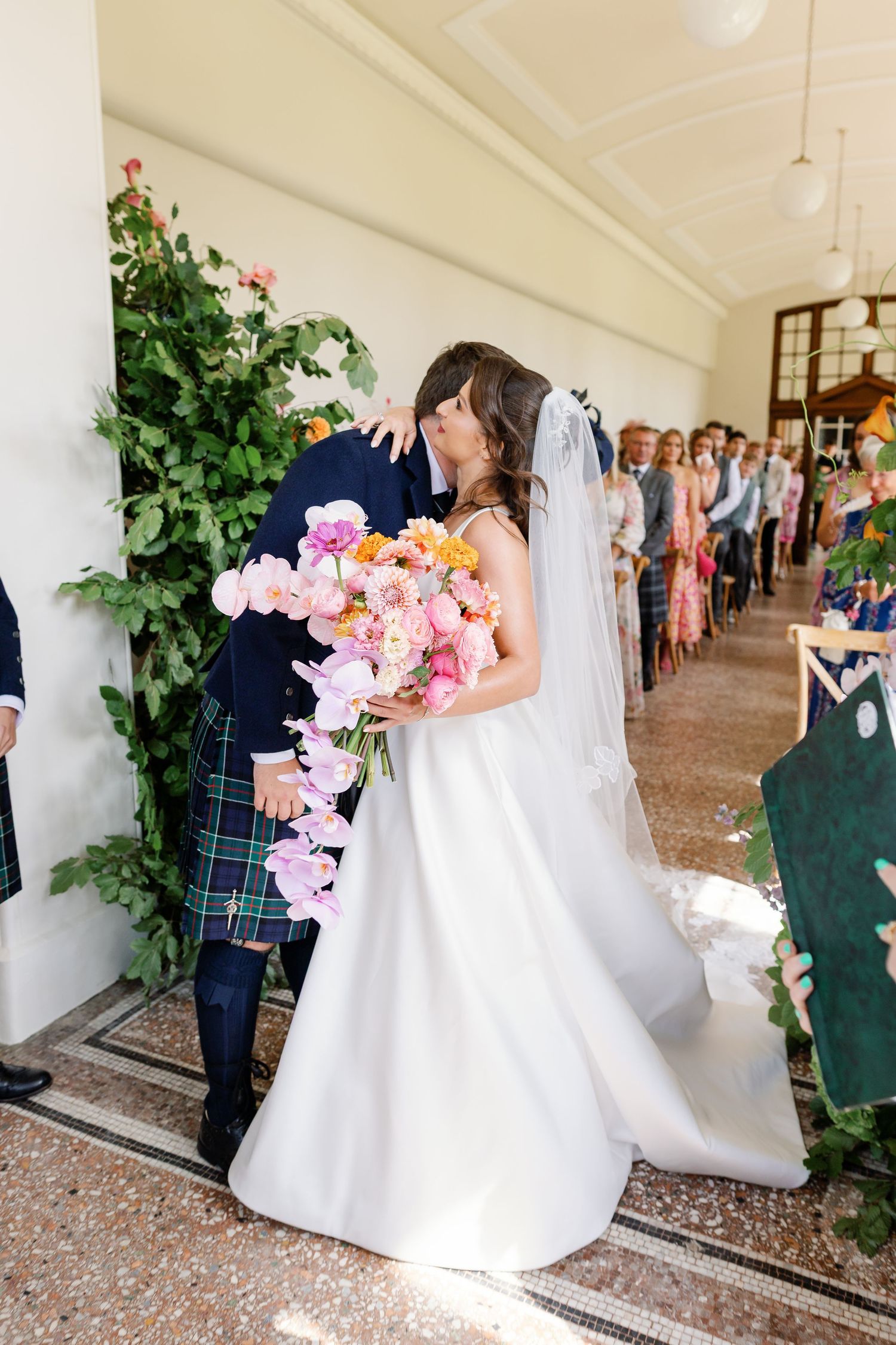 A wedding couple embraces in an elegant hallway with white columns and greenery during their first look moment.