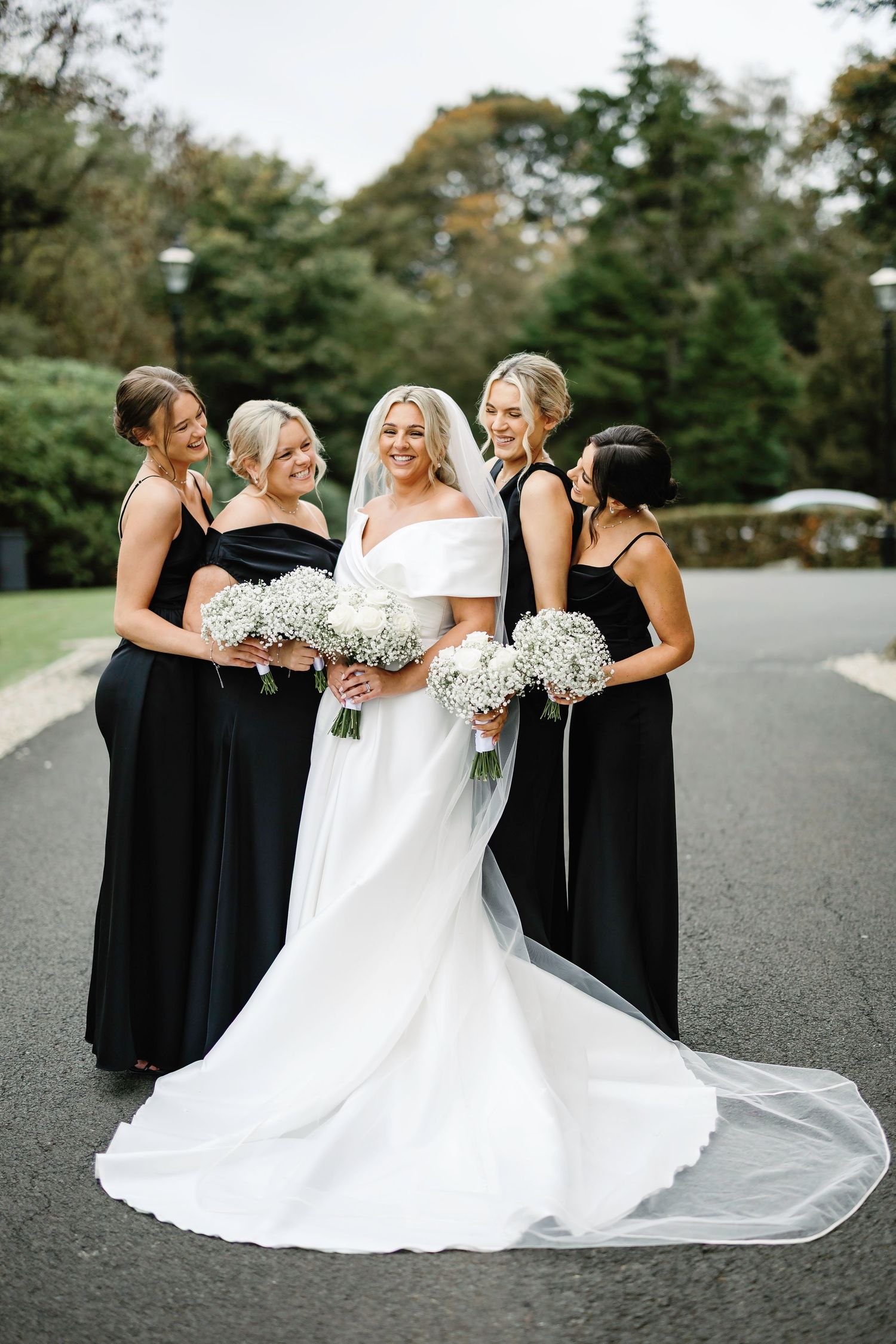 Bridal party in elegant black bridesmaid dresses and white wedding gown pose together holding baby's breath bouquets.