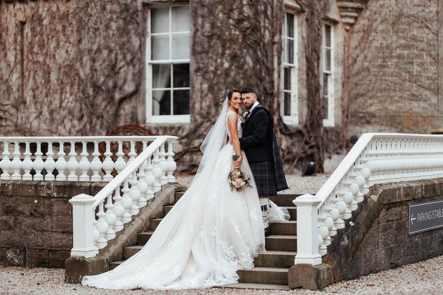 A couple in wedding attire pose elegantly on white stone stairs of a vine-covered historic building.
