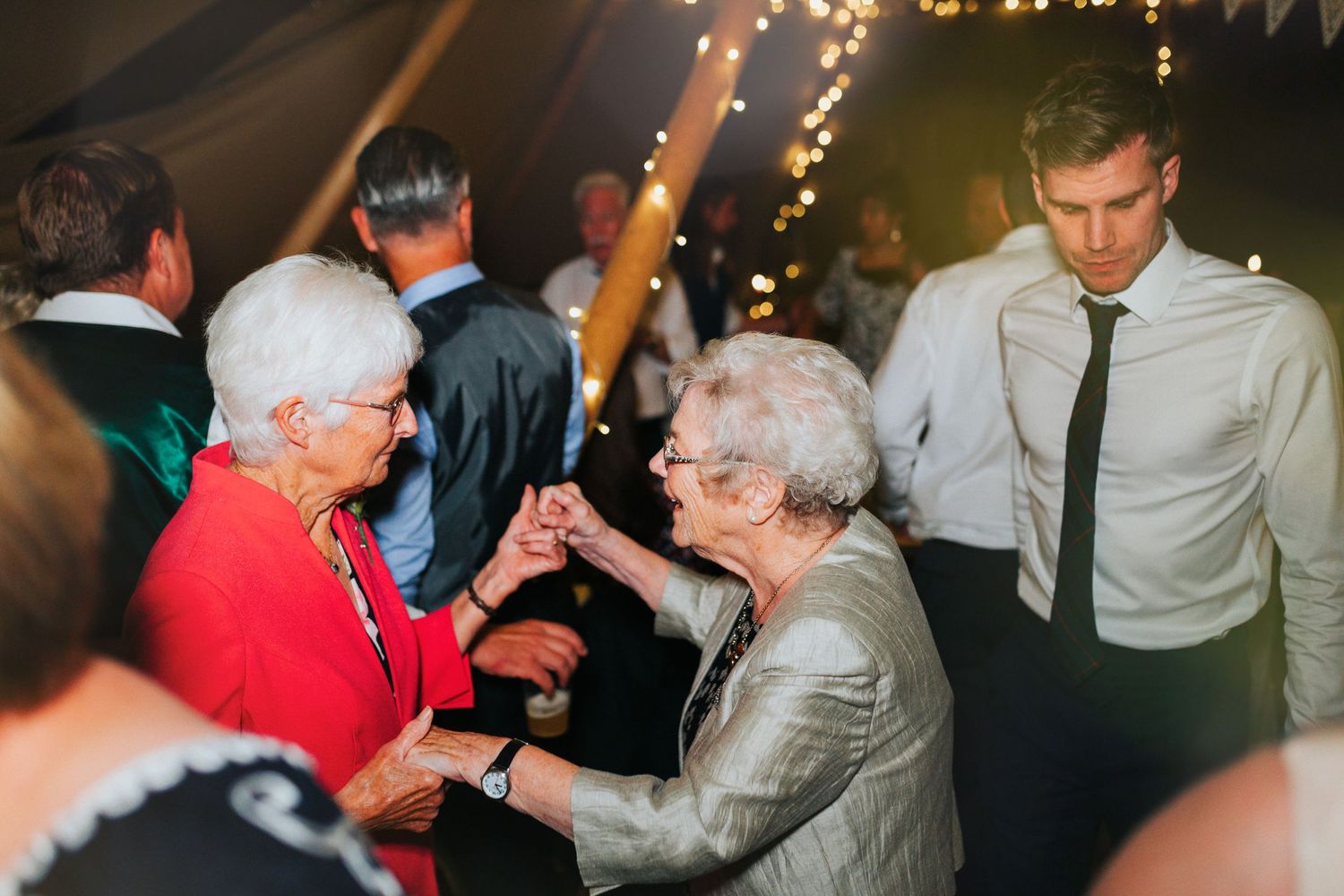 Two elderly ladies in festive spirits dance together at an evening celebration with string lights in the background.