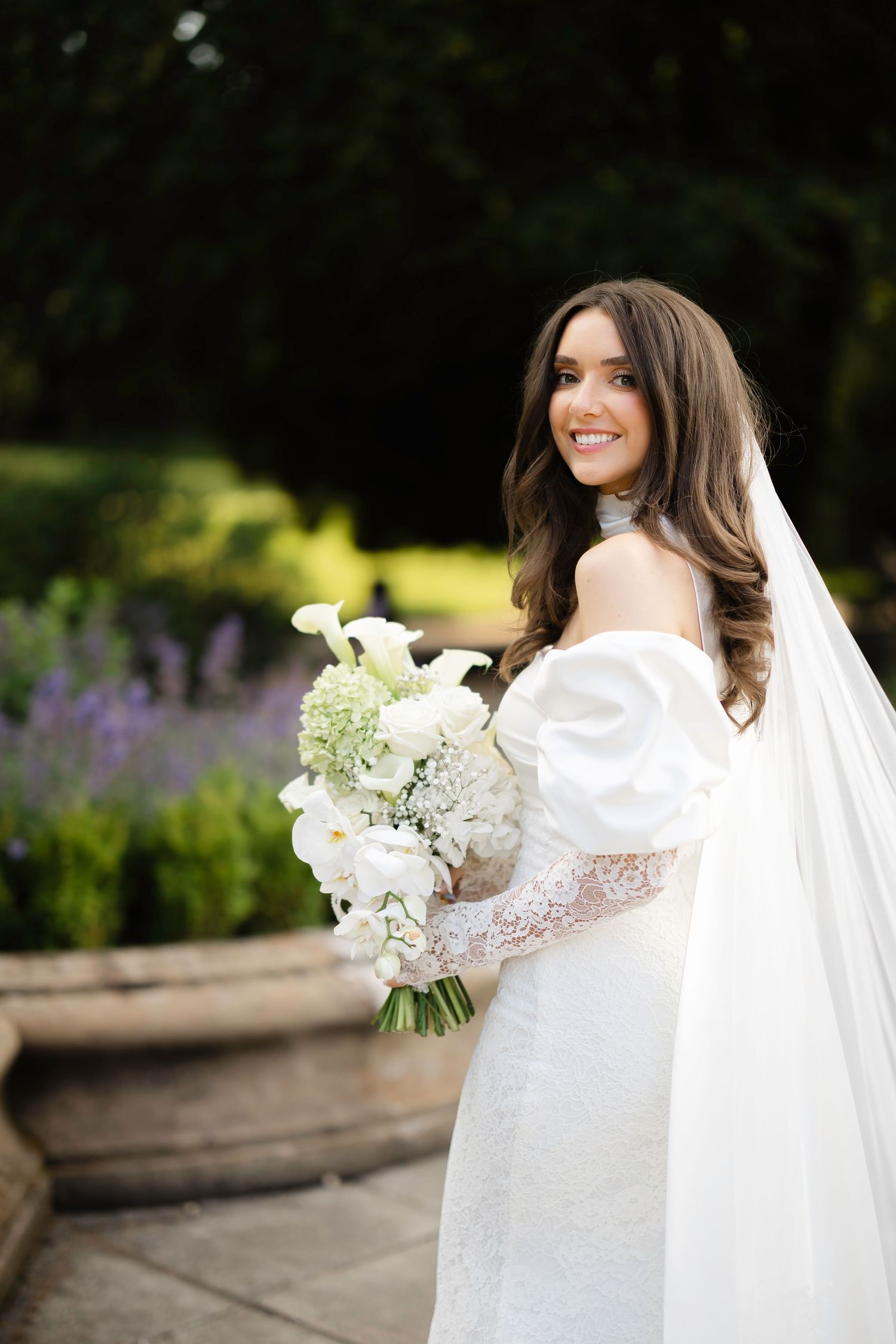 Smiling bride in white off-shoulder gown holds bouquet of white flowers in lush garden setting.