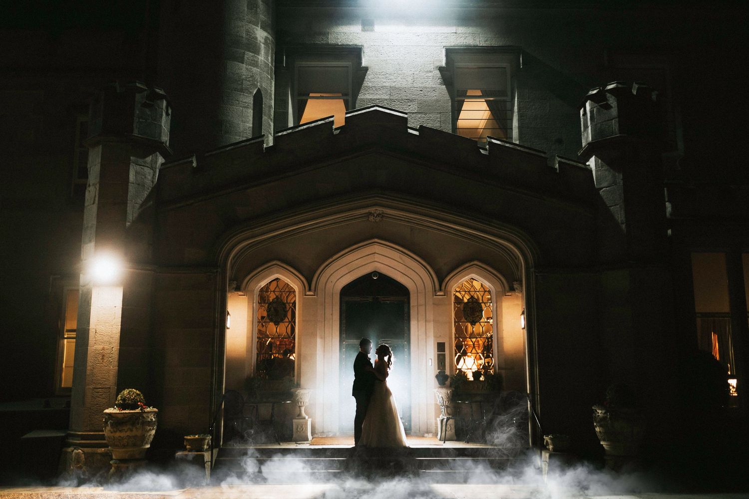 Silhouetted figures stand in a gothic archway entrance as smoke drifts across the illuminated stone doorway.
