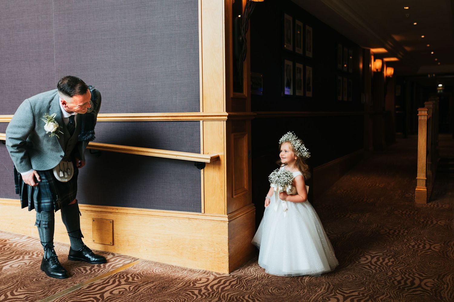 A person in blue leans against a wall while someone in a white dress stands around the corner in a dimly lit space.