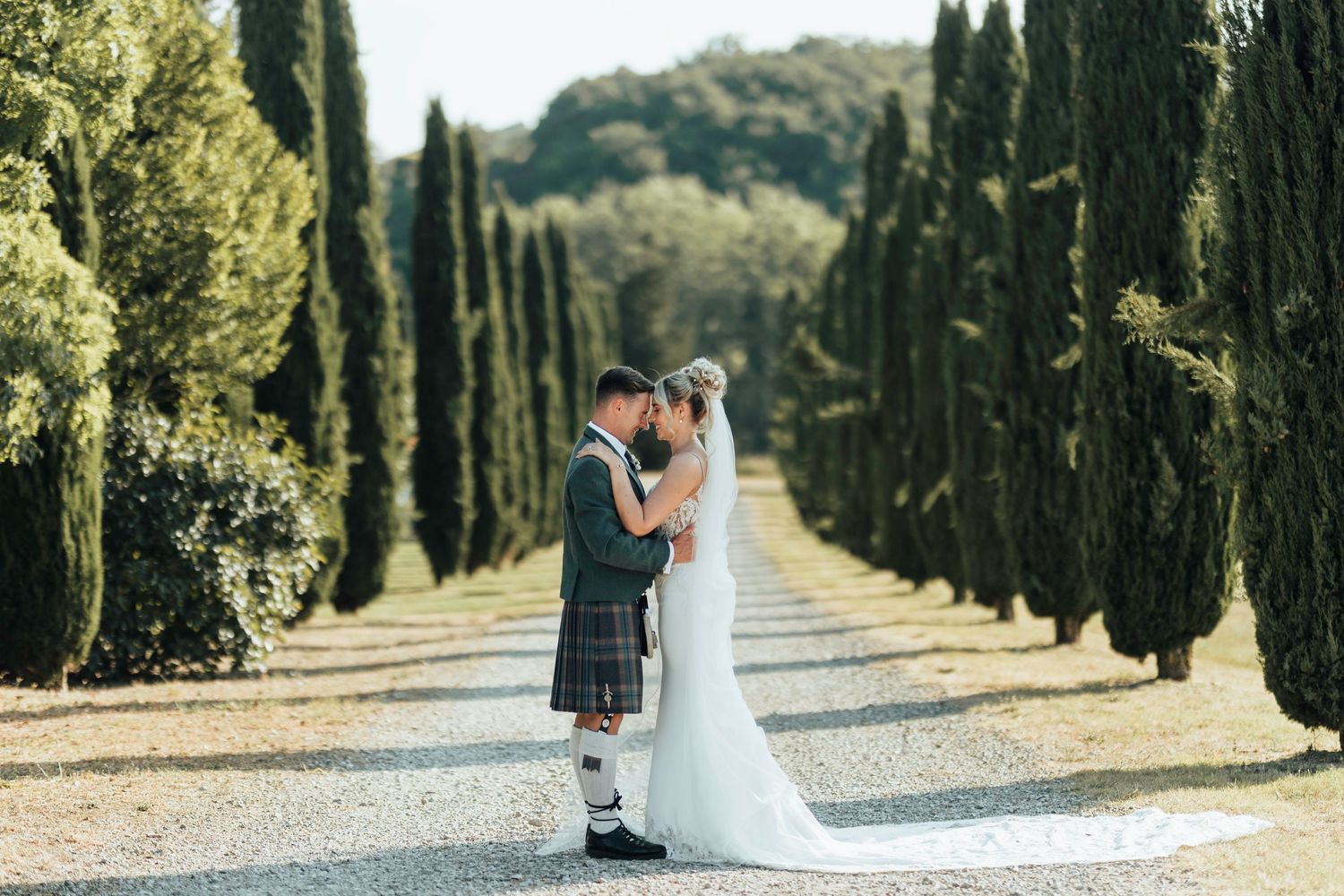 A romantic couple embraces on a cypress tree-lined path in a Tuscan garden setting.