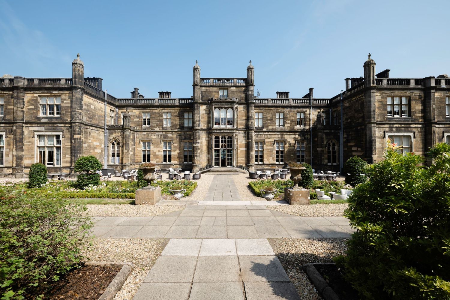 Grand stone manor house with manicured gardens, gravel pathways, and ornamental planters under a clear blue sky.