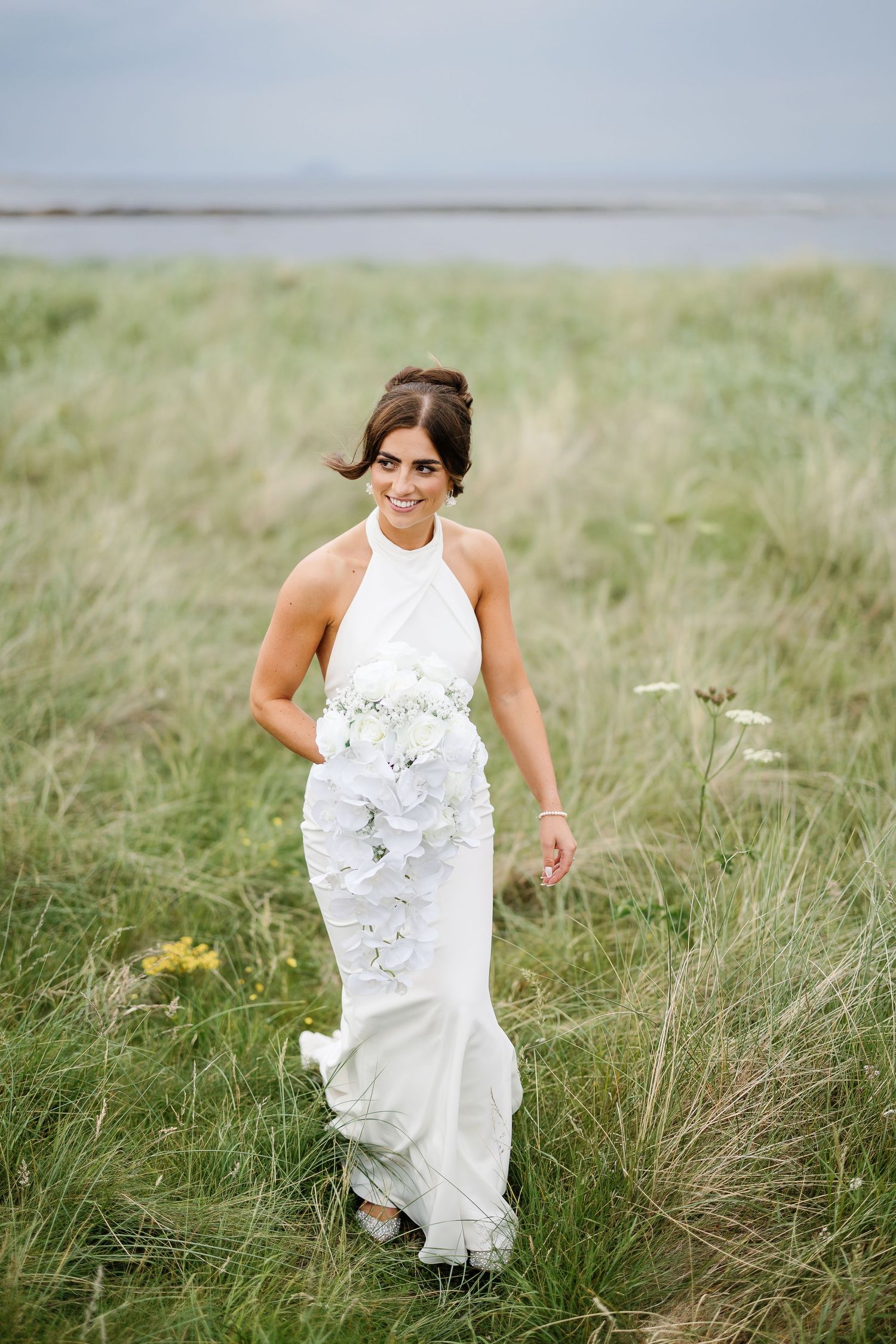 A person in a white sleeveless wedding dress runs playfully through tall grass in a field on a sunny day.