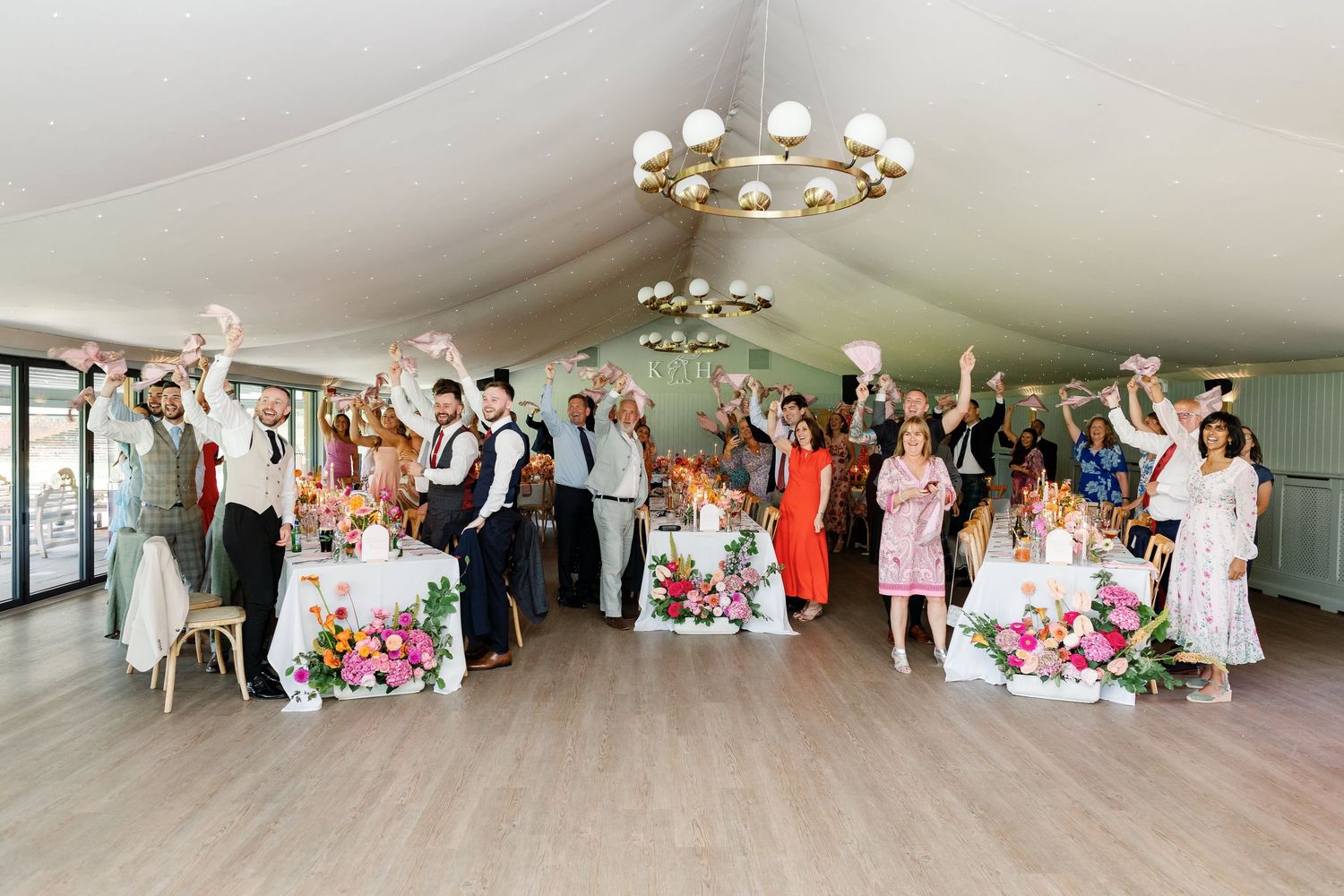 Wedding guests celebrate and dance together inside an elegant white tent with floral decorations and chandeliers.
