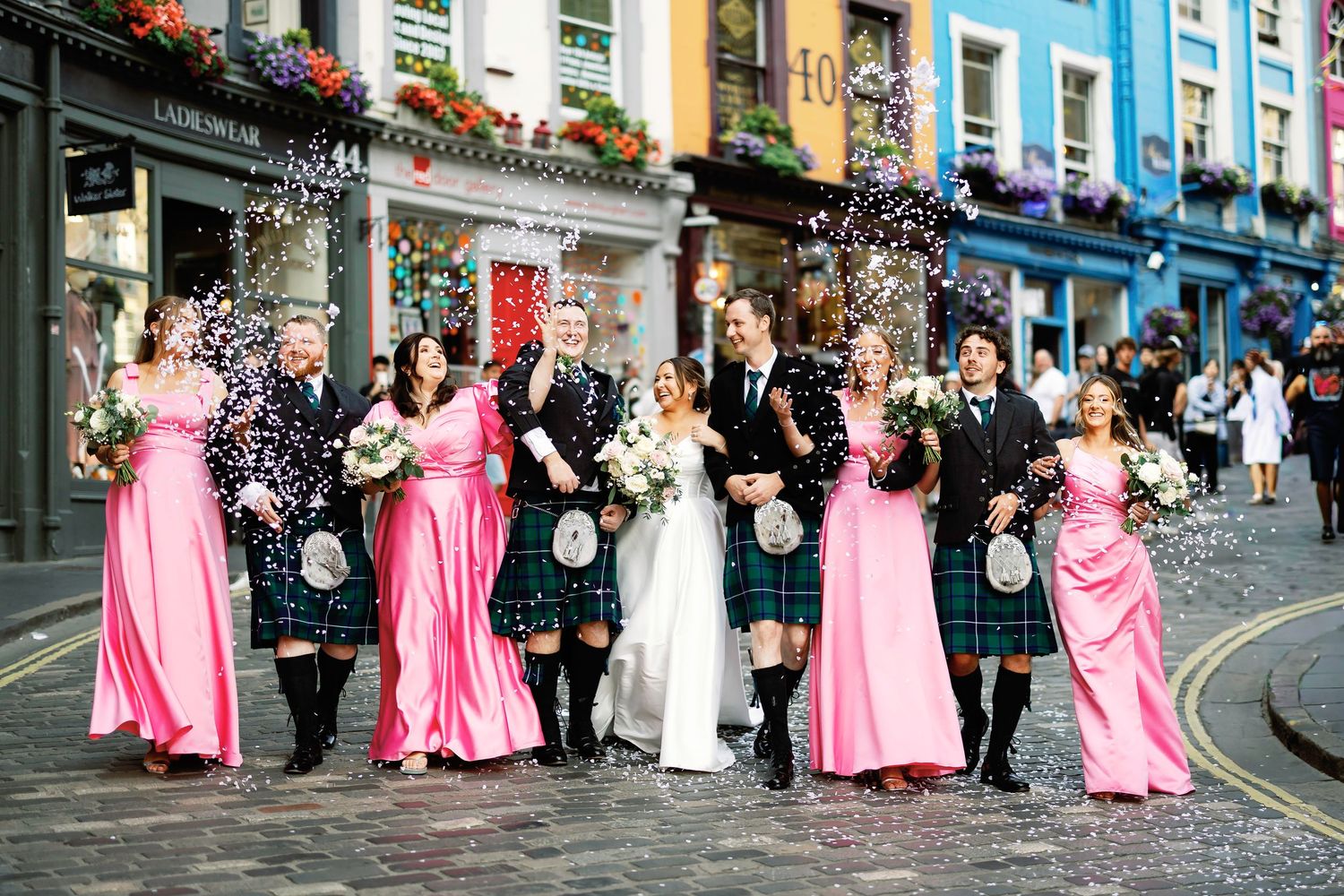 Wedding party in pink and black attire poses on a charming cobblestone street with colorful buildings in the background.