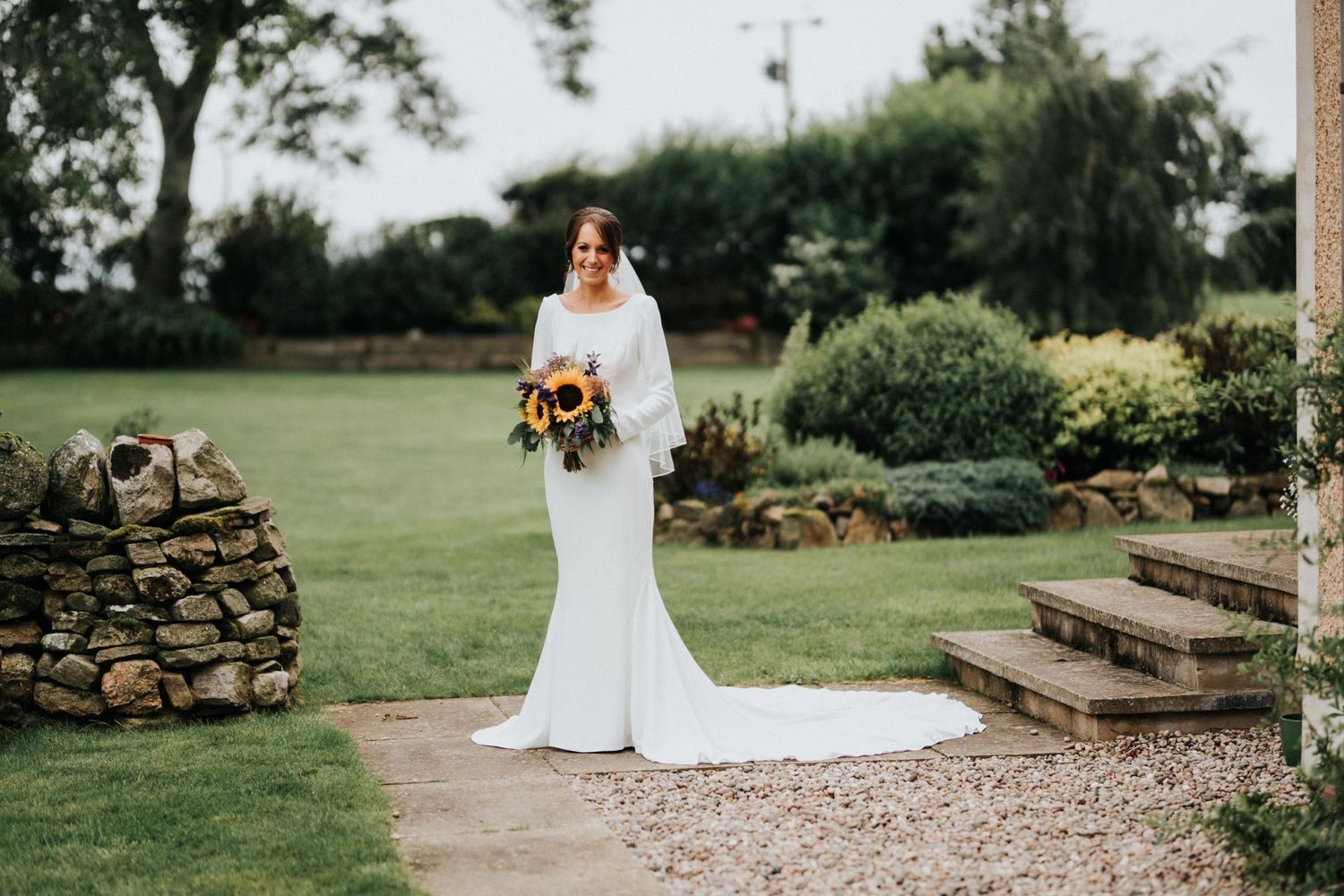 A bride in a fitted white gown with a long train holds a vibrant autumn bouquet while standing in a garden setting.