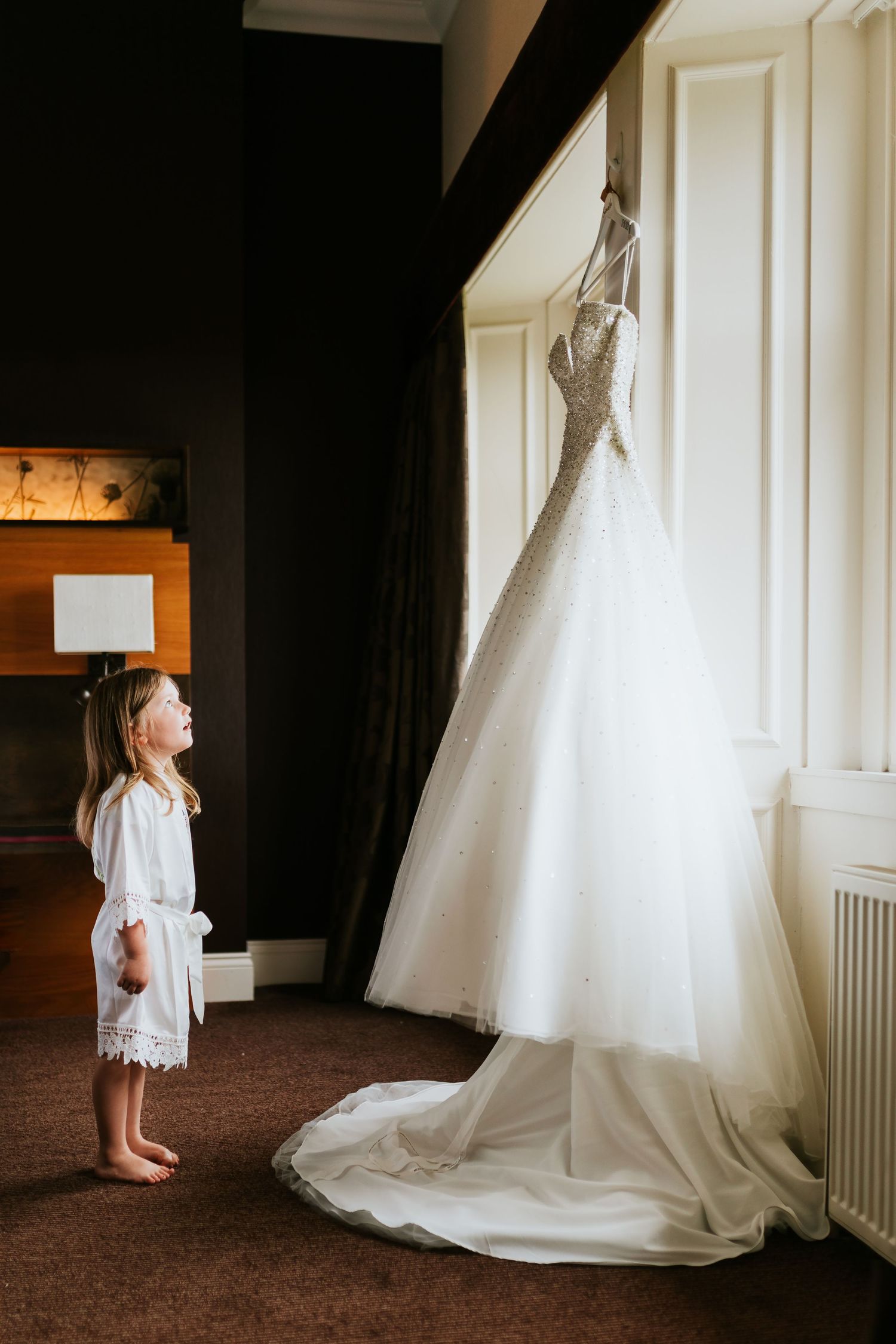 Child in white lace dress looks up at elegant wedding gown hanging by window in dramatic lighting.