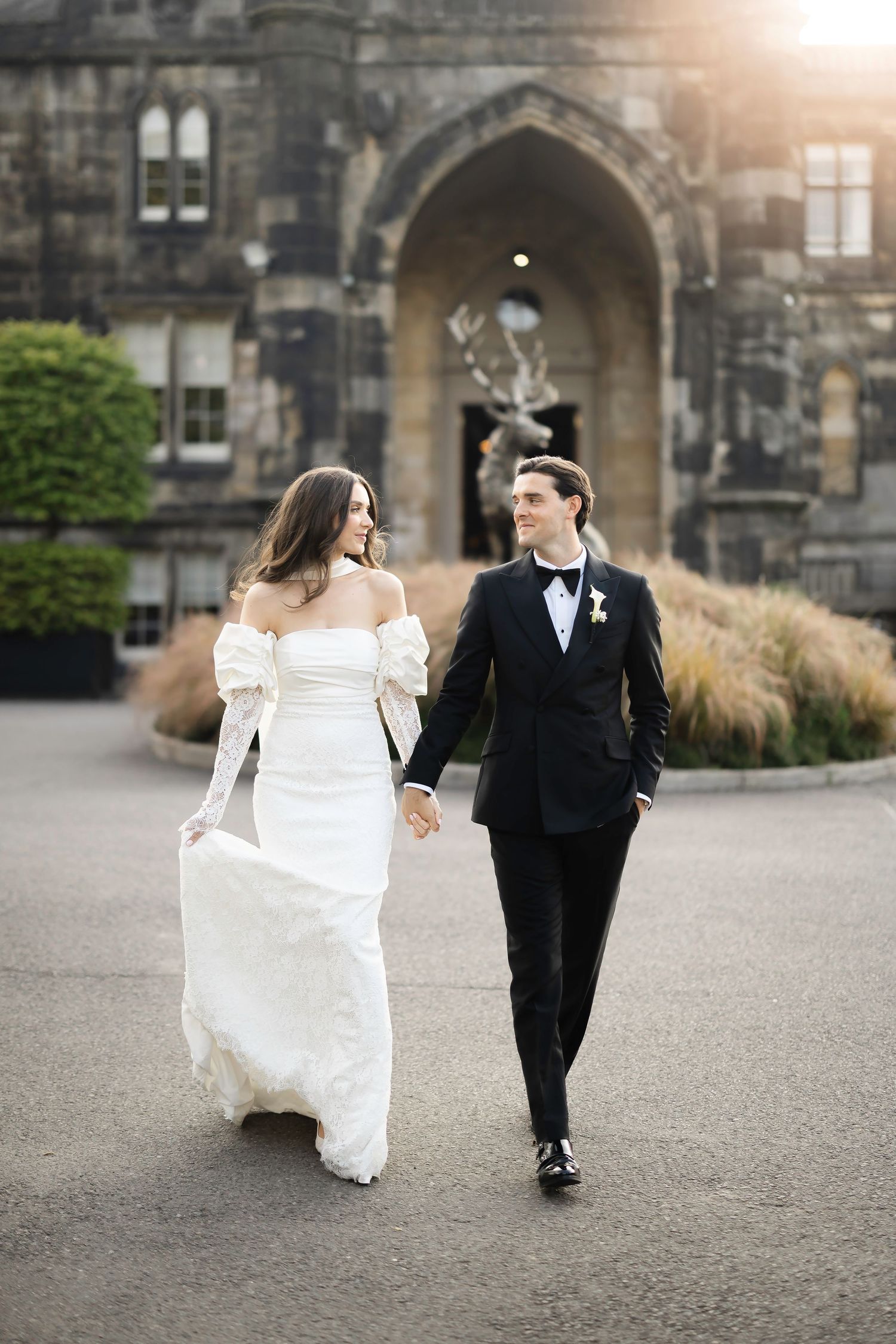 Bride in white off-shoulder gown and groom in black tuxedo walking hand-in-hand before a grand stone building.
