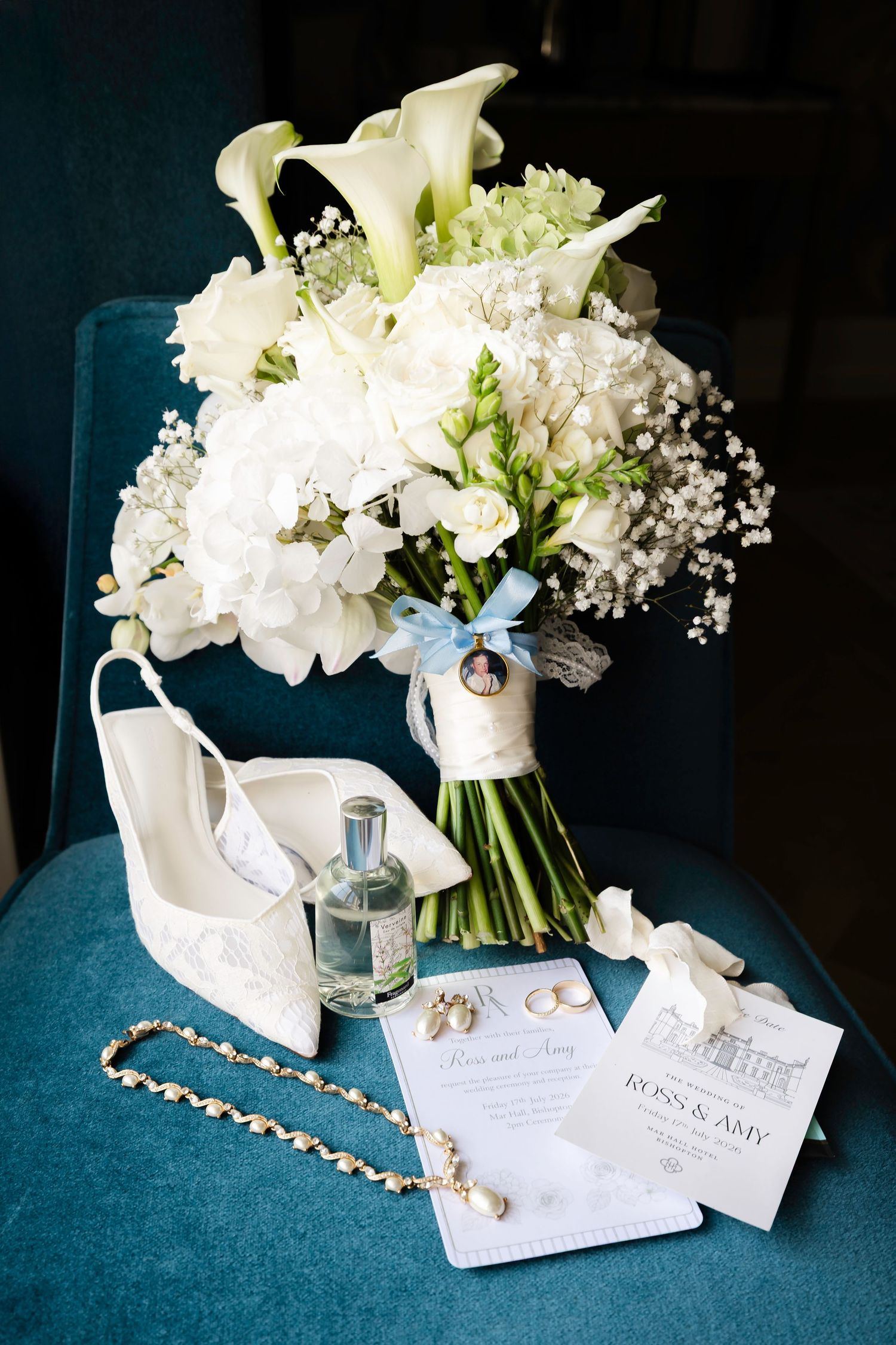 Elegant wedding flat lay with white calla lily bouquet, bridal heels, perfume, jewelry, and Ross & Amy wedding invitations on teal chair.