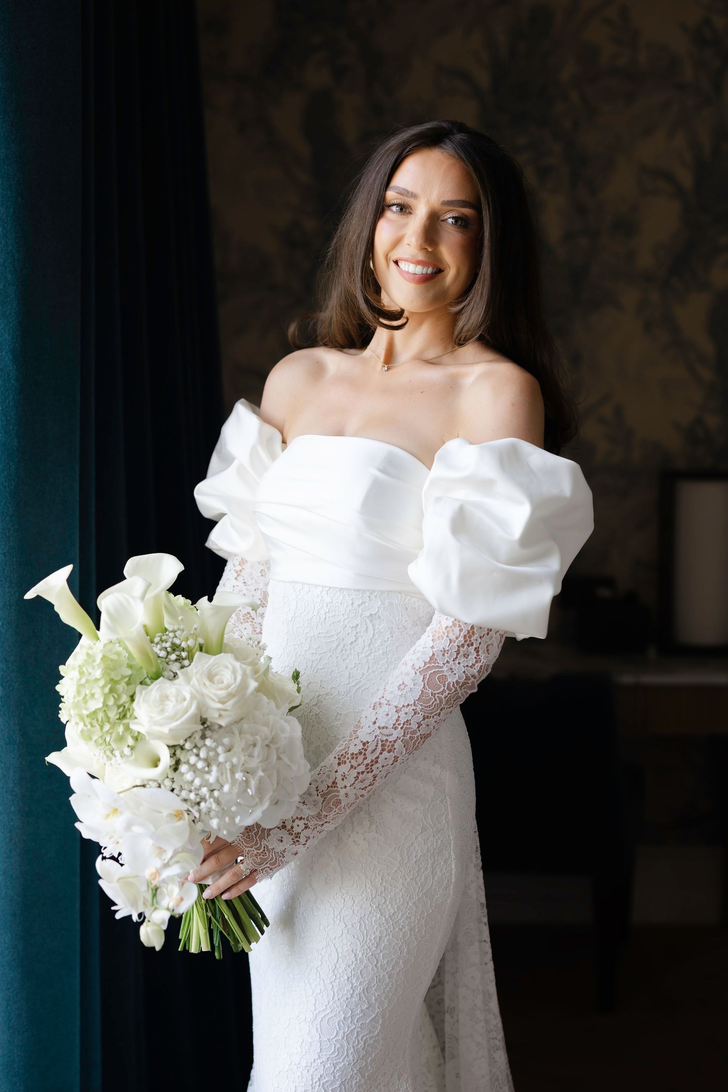 Smiling bride in off-shoulder white lace gown holding elegant white bouquet of roses and calla lilies indoors.