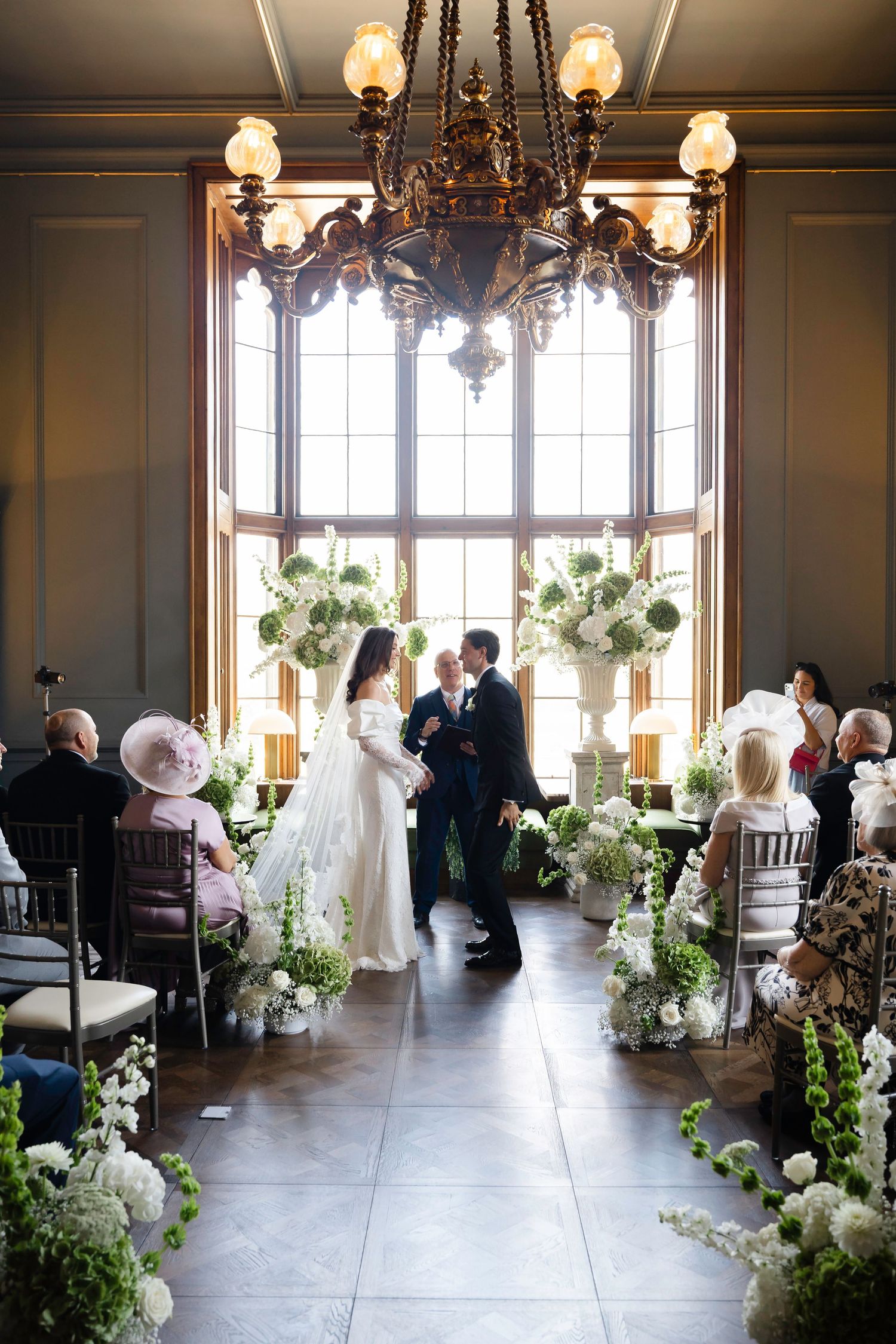 Elegant indoor wedding ceremony with couple at altar, white floral arrangements, chandelier, and seated guests in a grand venue.