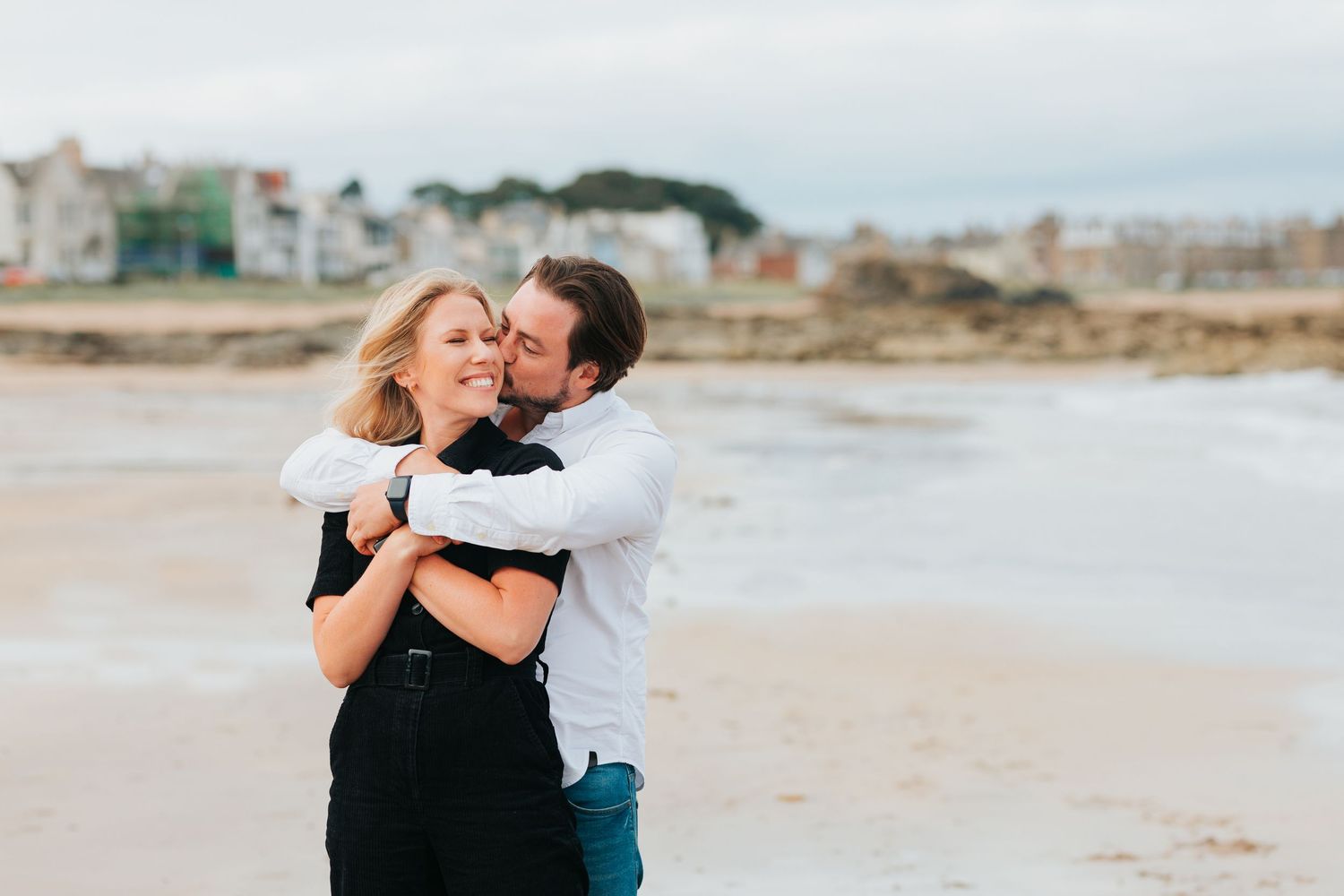 A romantic couple embraces and laughs together on a cloudy beach with buildings in the background.