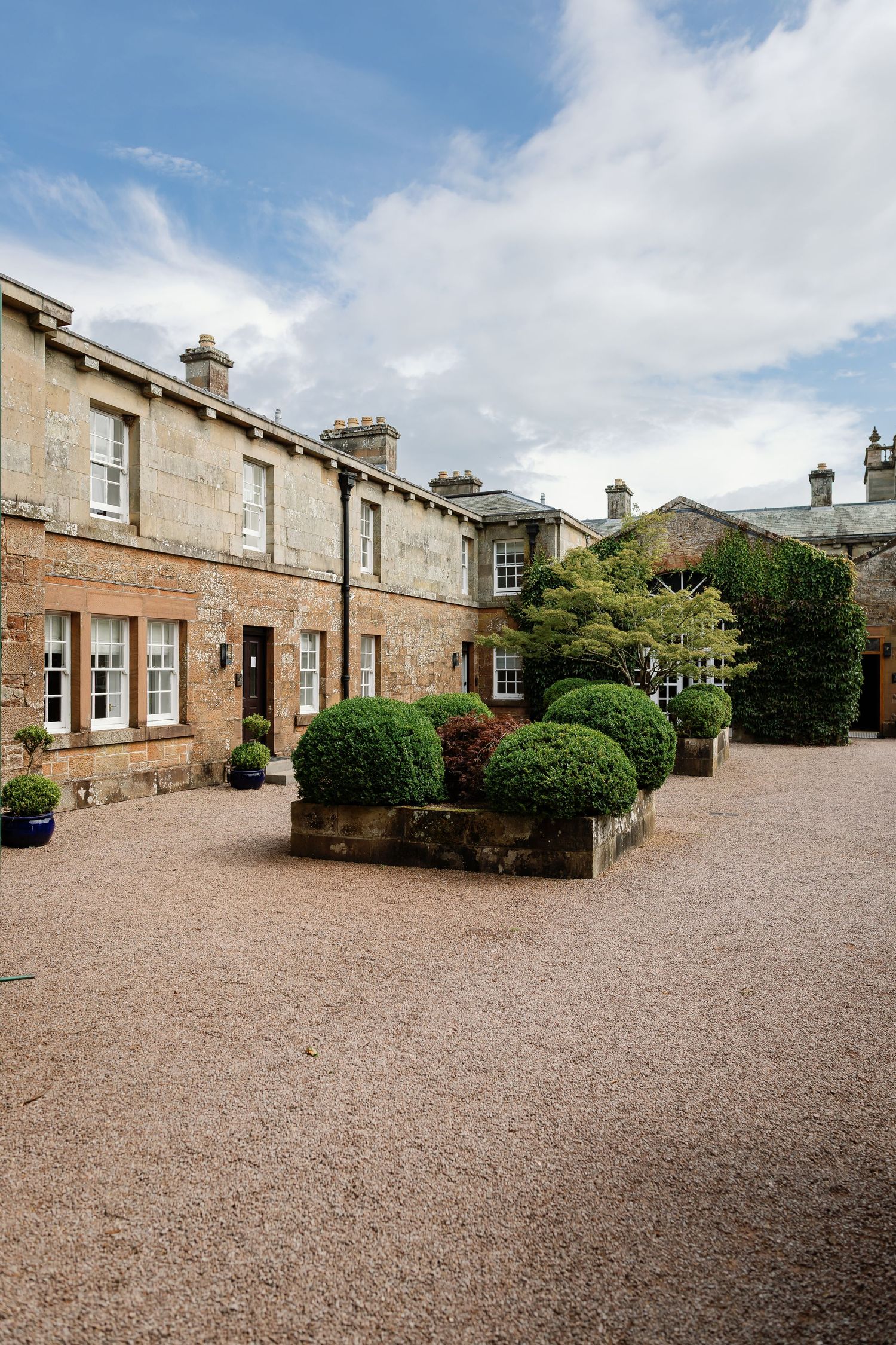 Historic stone courtyard with gravel driveway surrounded by traditional Scottish architecture and manicured shrubs.