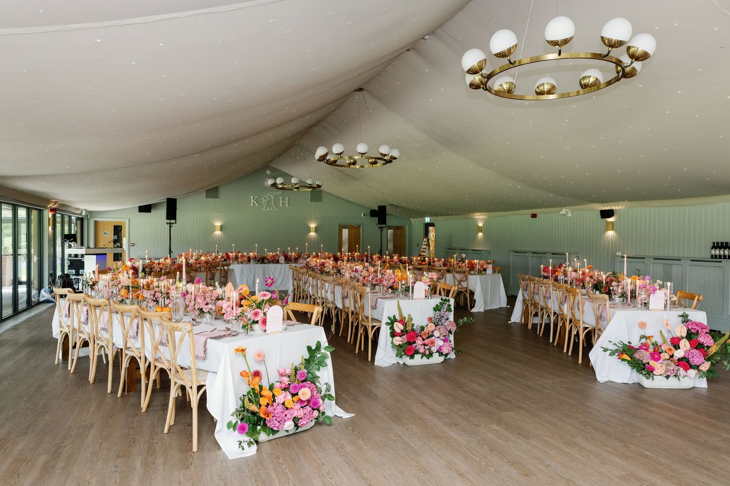 A large reception hall decorated with white tables, pink floral centerpieces, and elegant chandeliers for a formal event.