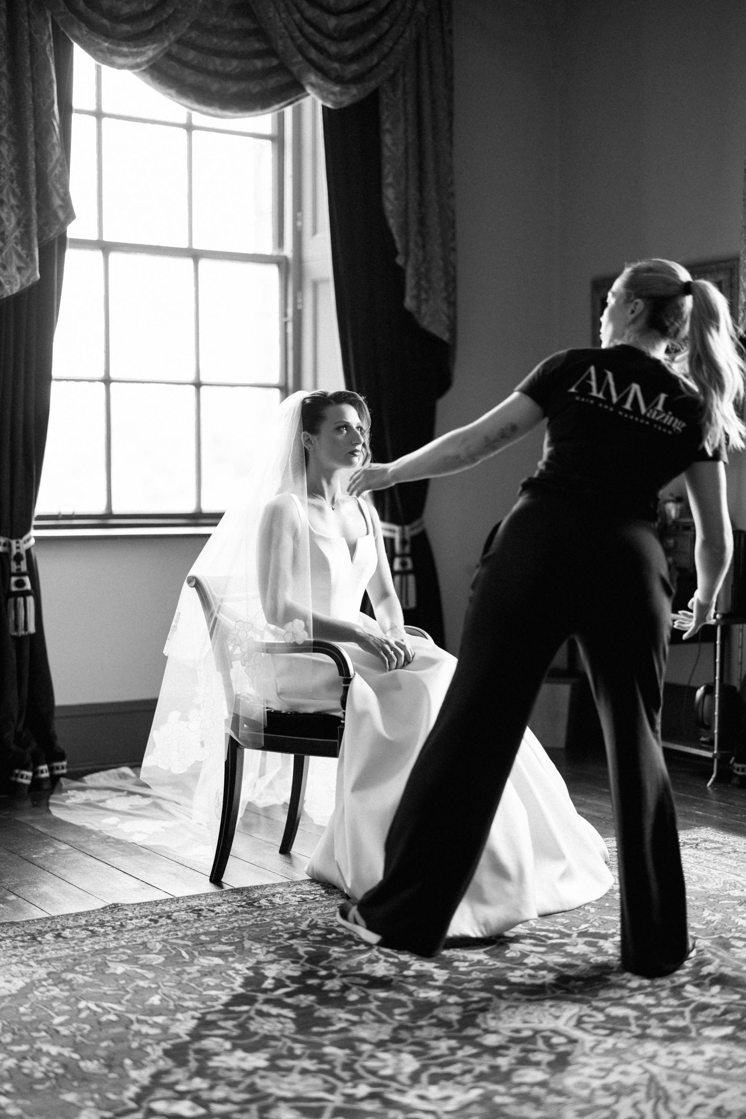 Black and white photo showing wedding preparation scene with a bride in white dress seated while stylist works on final touches.