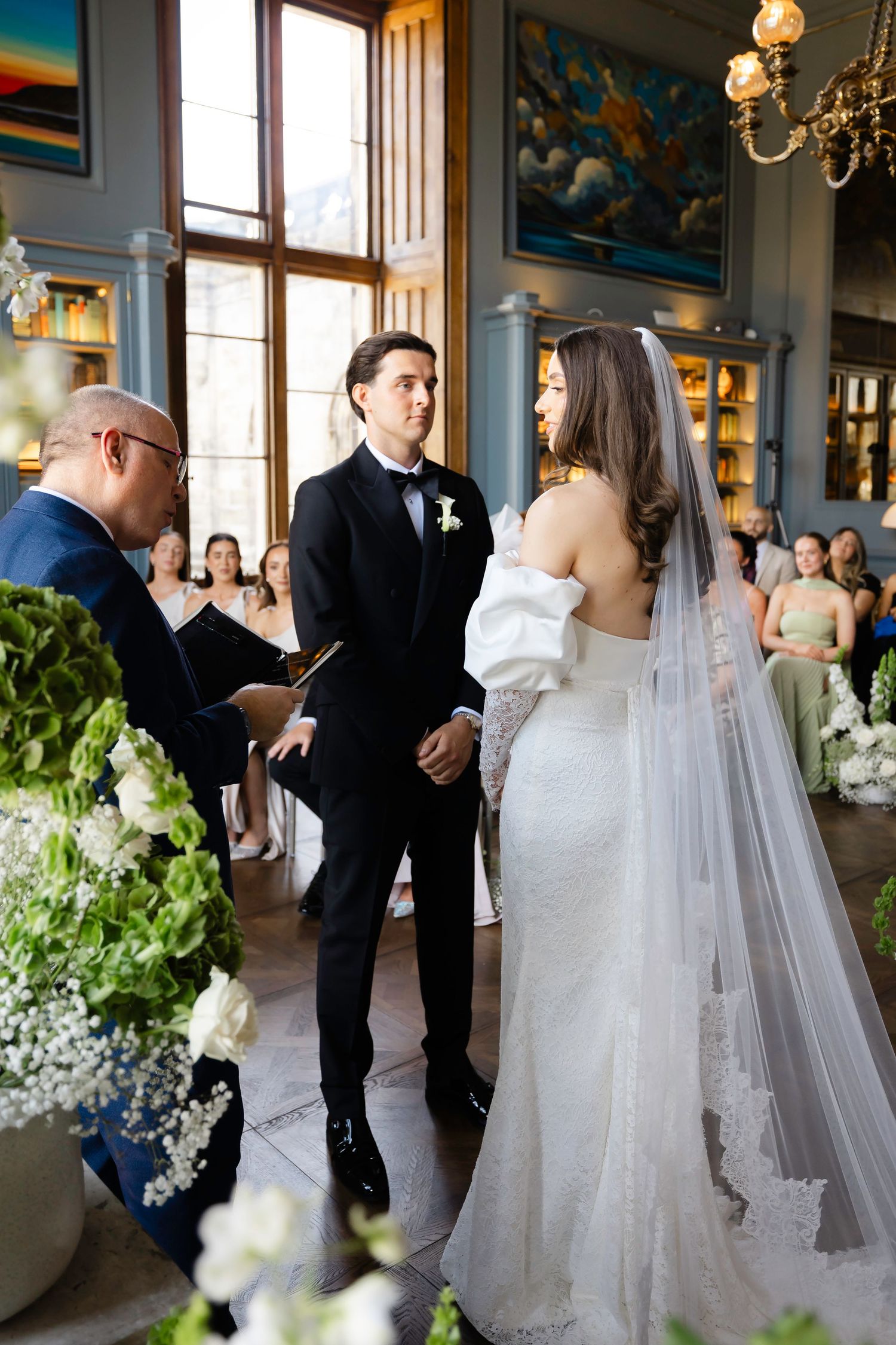 Bride and groom exchange vows at elegant indoor wedding ceremony with white floral arrangements and large windows.