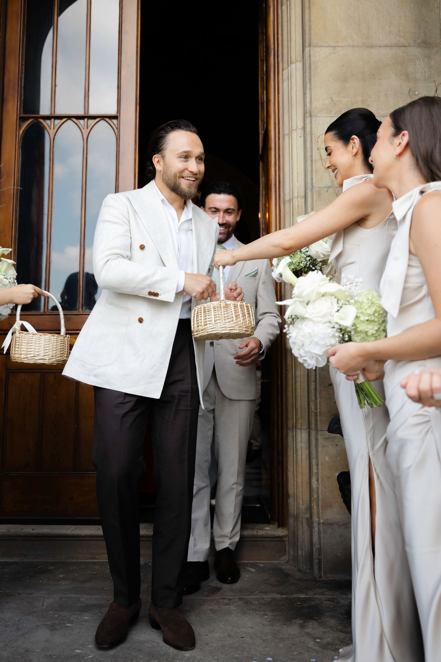 Smiling groom in white jacket greets bridesmaids holding flowers and basket outside church doors.