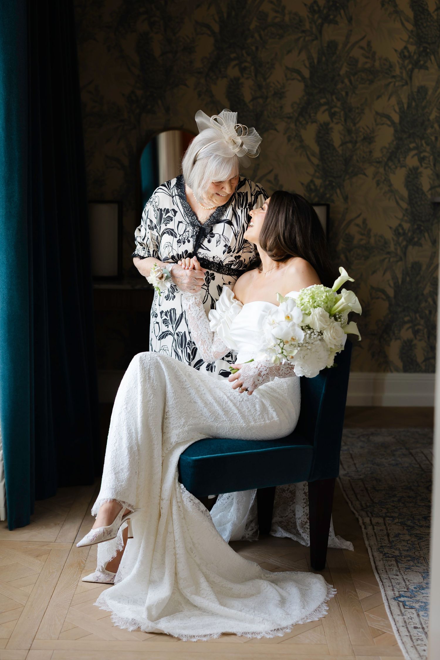 Bride in white lace gown holds bouquet while elderly woman in black lace dress shares tender moment before wedding.