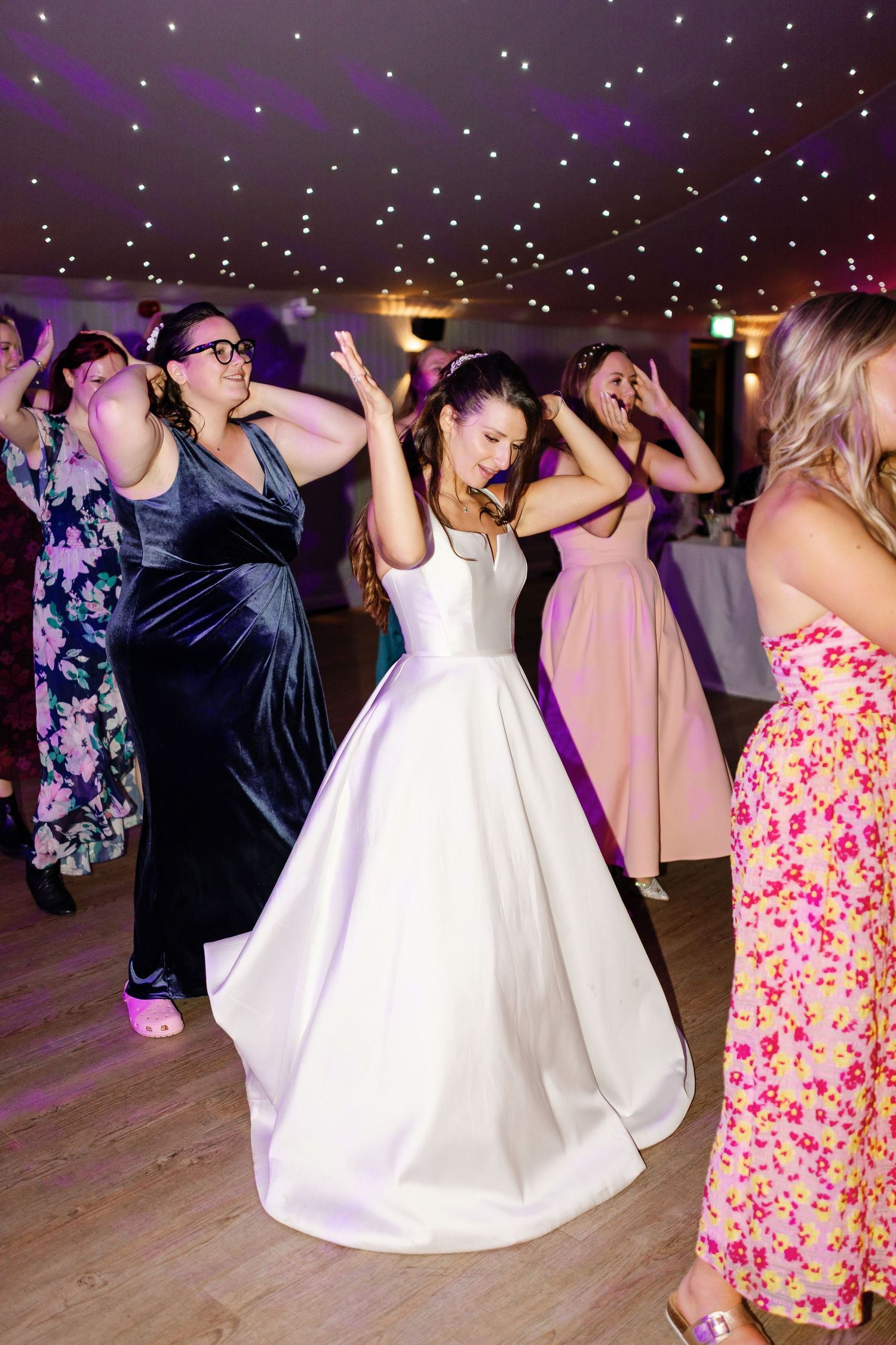 Wedding guests dance and celebrate on the dance floor at a reception under twinkling lights.