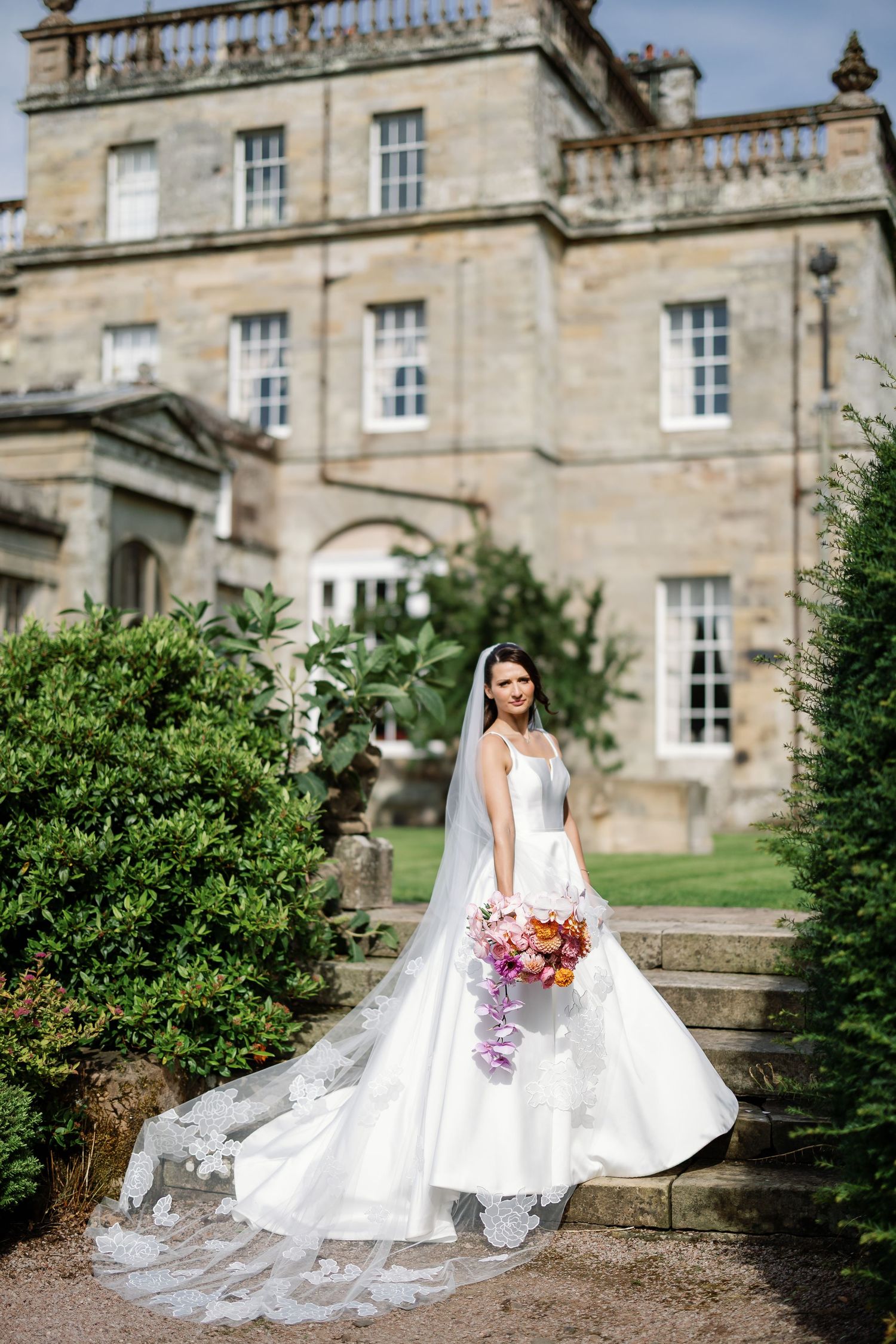 A bride in an elegant white gown poses in front of a grand historic manor house with stone architecture.