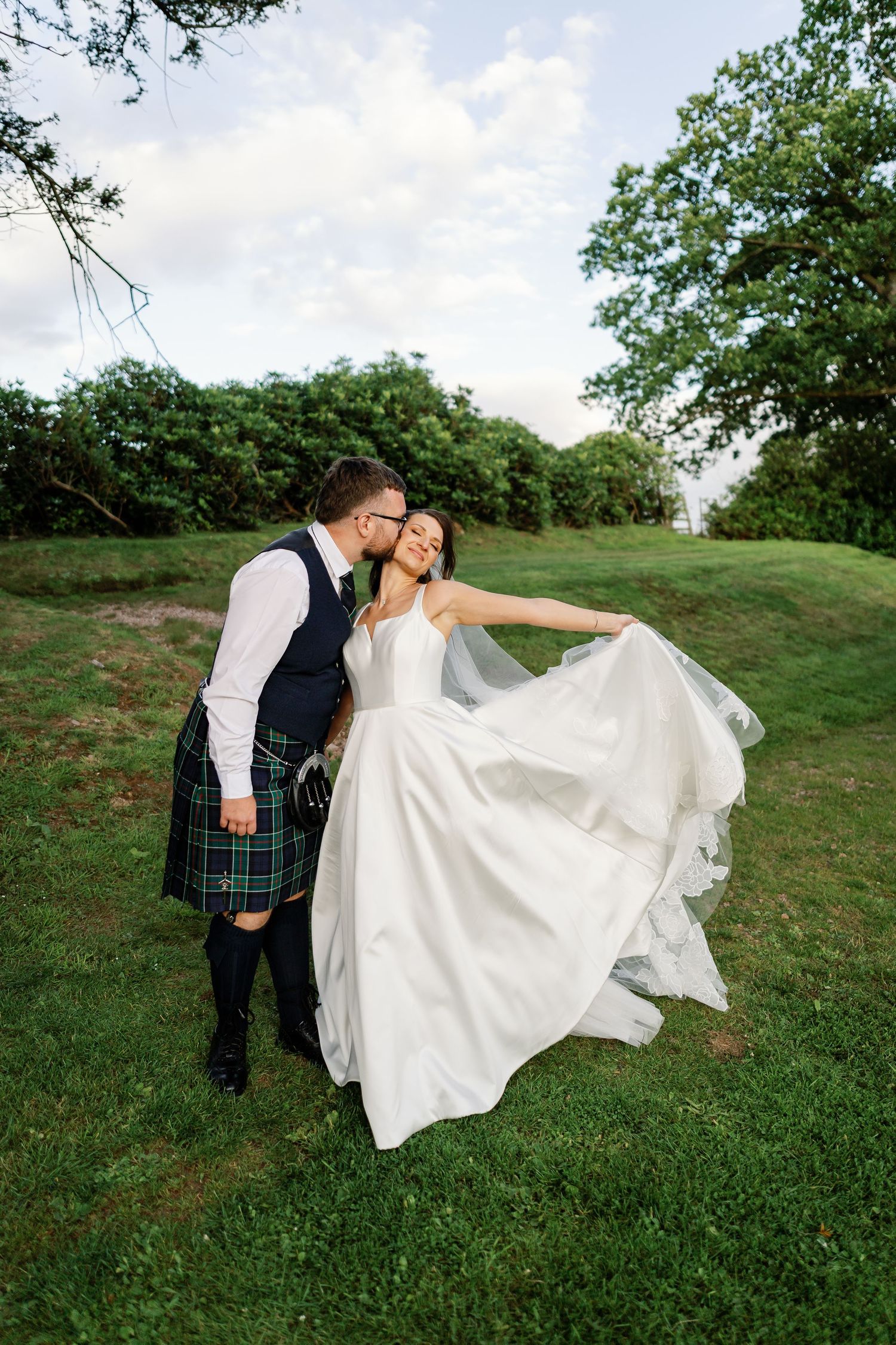 A couple in wedding attire poses playfully on a grassy hillside with the bride's dress flowing in the wind.
