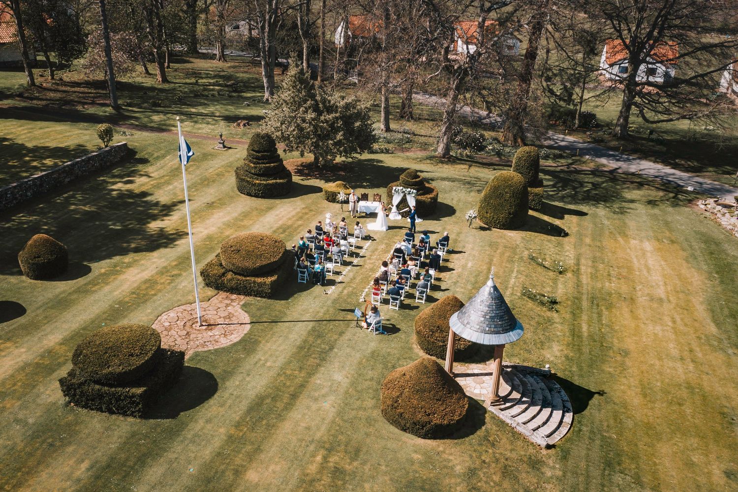 Aerial view of a garden party setup with white tents, tables, and chairs arranged on a manicured lawn with decorative shrubs.
