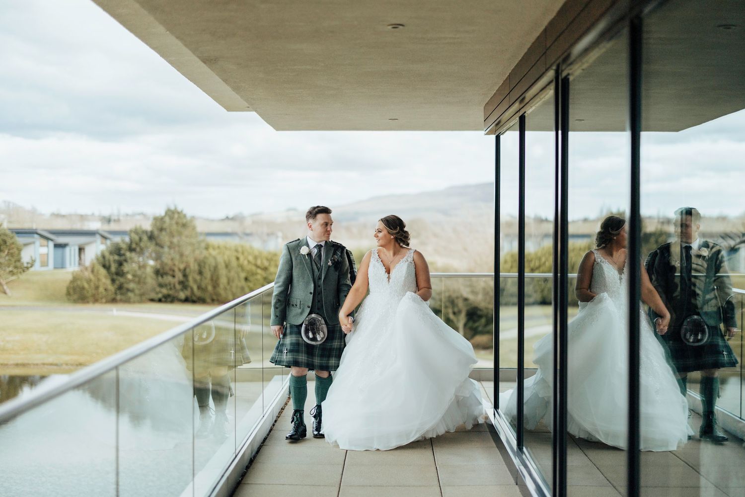 A couple in wedding attire stands on a modern balcony overlooking scenic mountain landscapes on their special day.