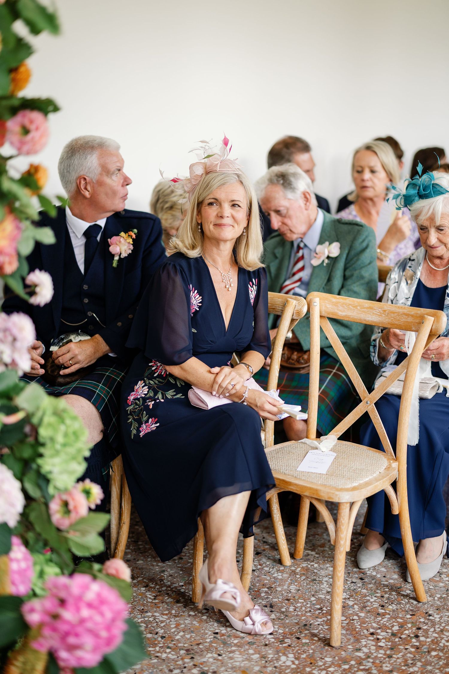 Wedding guests seated in wooden cross-back chairs at an outdoor ceremony with pink floral decorations.
