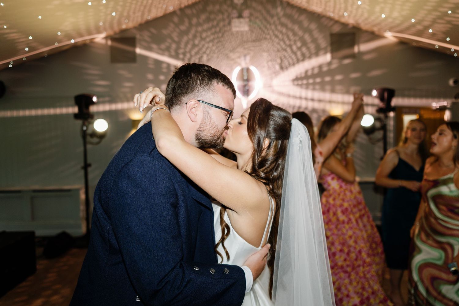 A couple shares a romantic kiss during their wedding reception under twinkling lights.