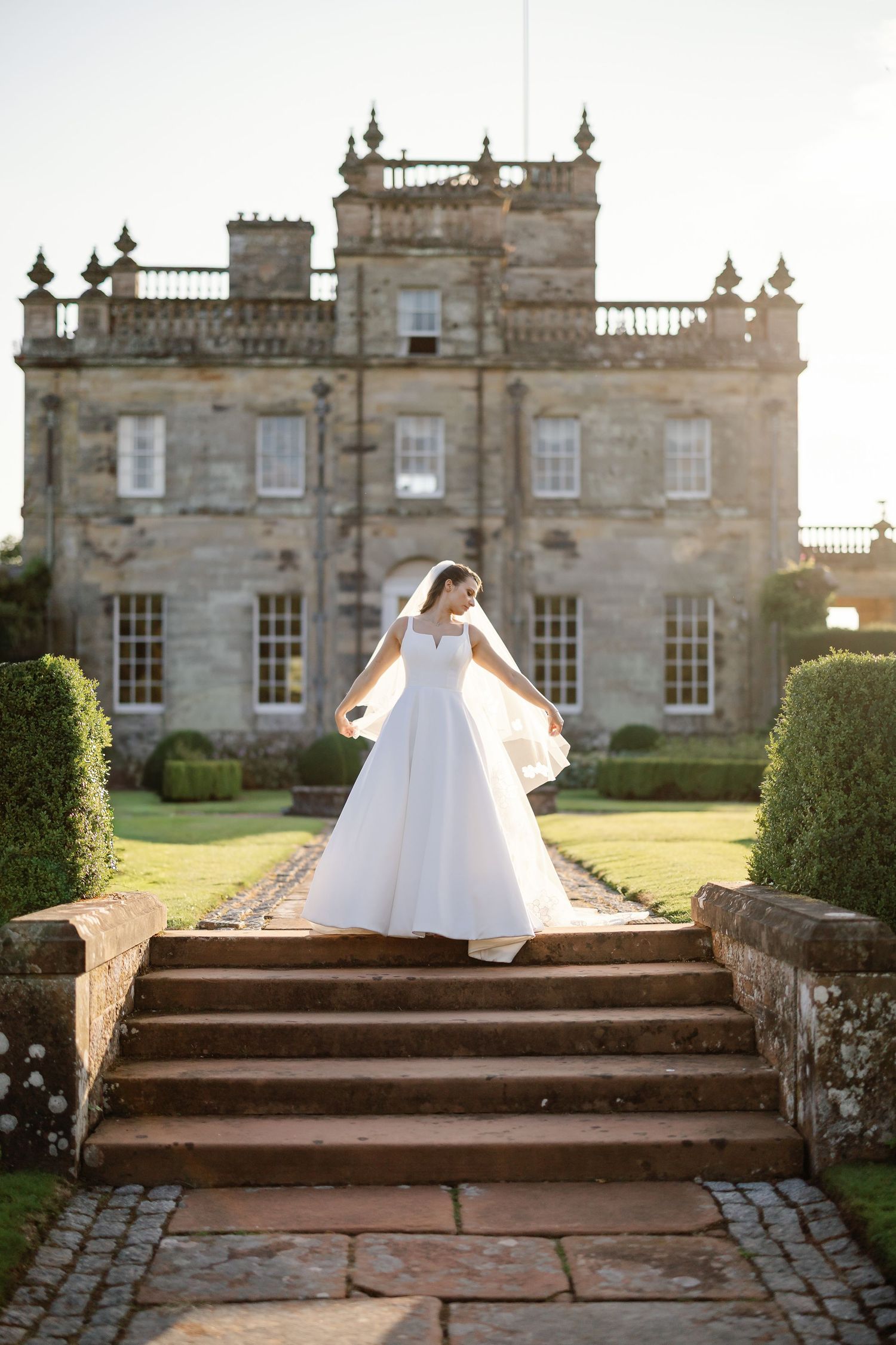 A white wedding dress is displayed on stone steps leading to a grand historic castle estate with manicured gardens.