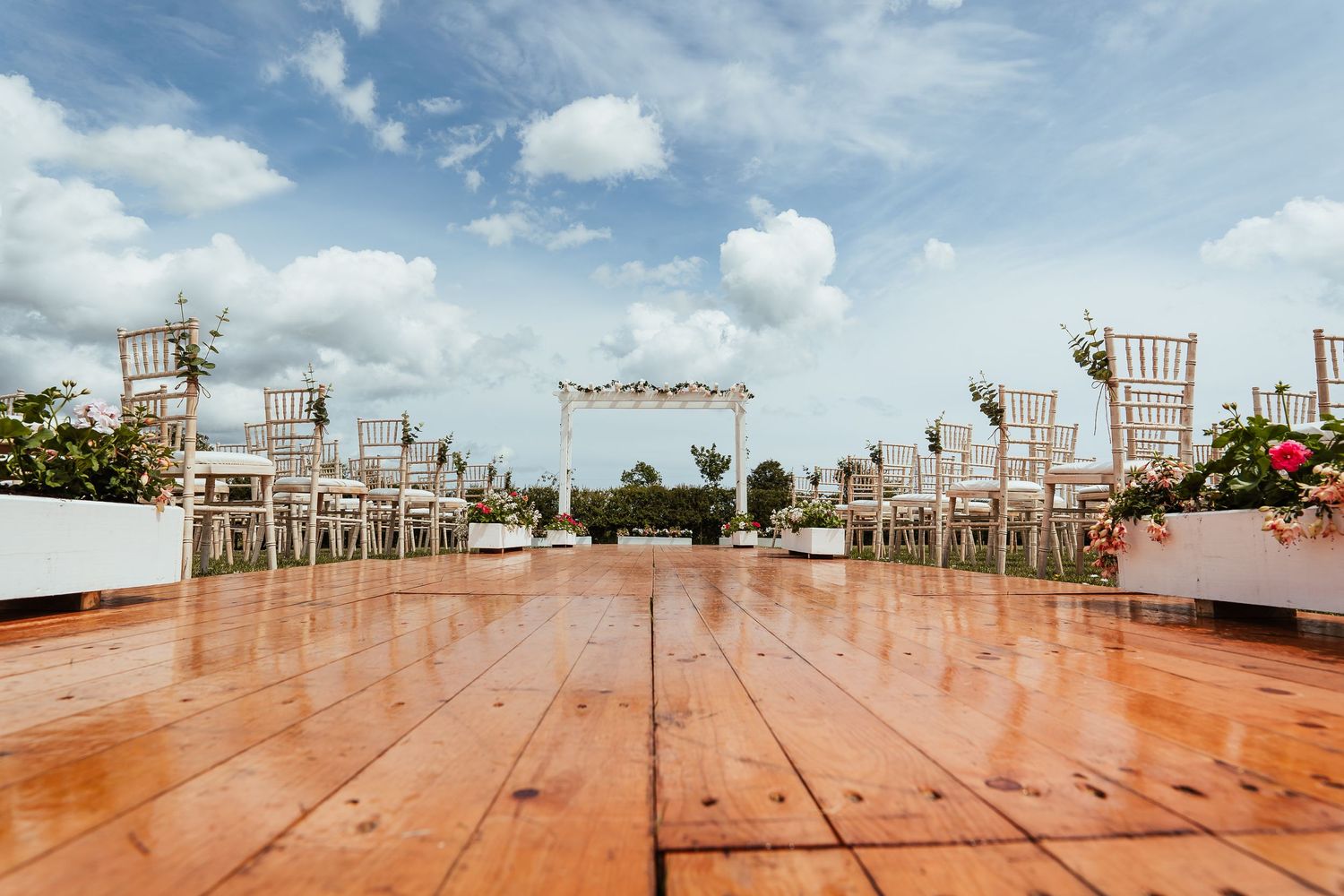 An outdoor wedding ceremony setup with wooden flooring and white chairs arranged in rows under a blue sky with fluffy clouds.