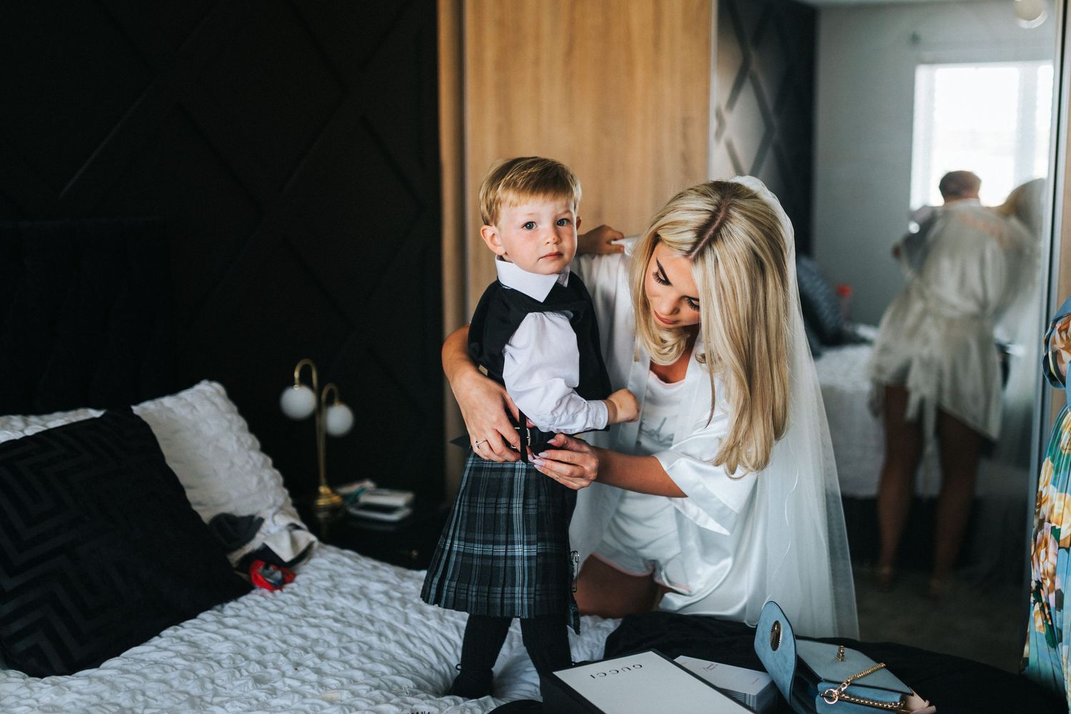 A boy wearing a kilt and bow tie gets dressed with assistance in a bedroom setting.