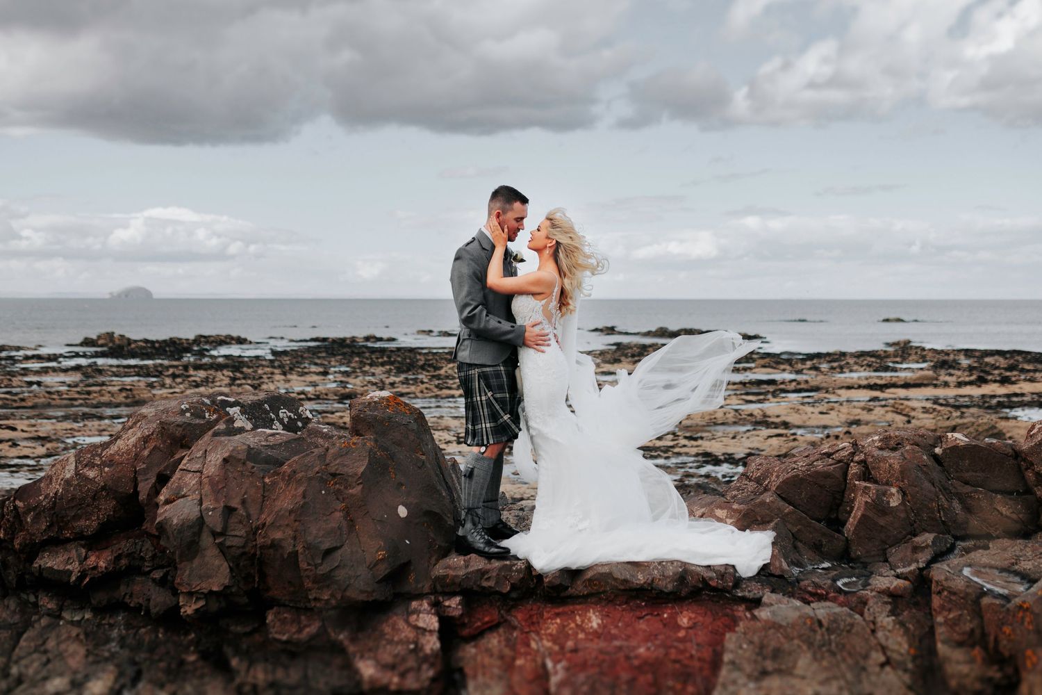 Couple embraces on rocky coastal shoreline as white wedding dress flows in the wind against dramatic cloudy sky.