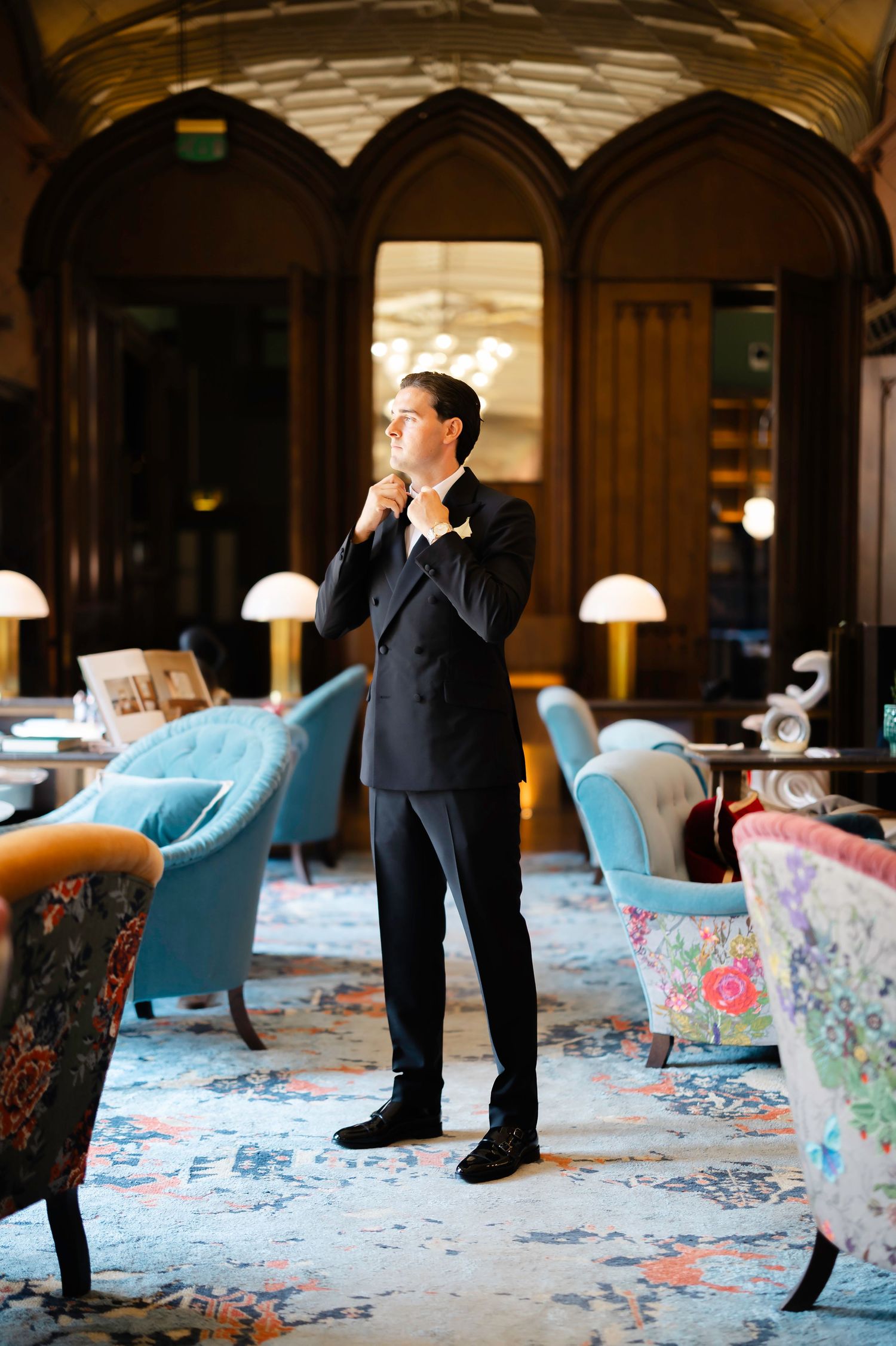 Groom in black tuxedo adjusting tie in elegant hotel lobby with blue velvet chairs and ornate decor.