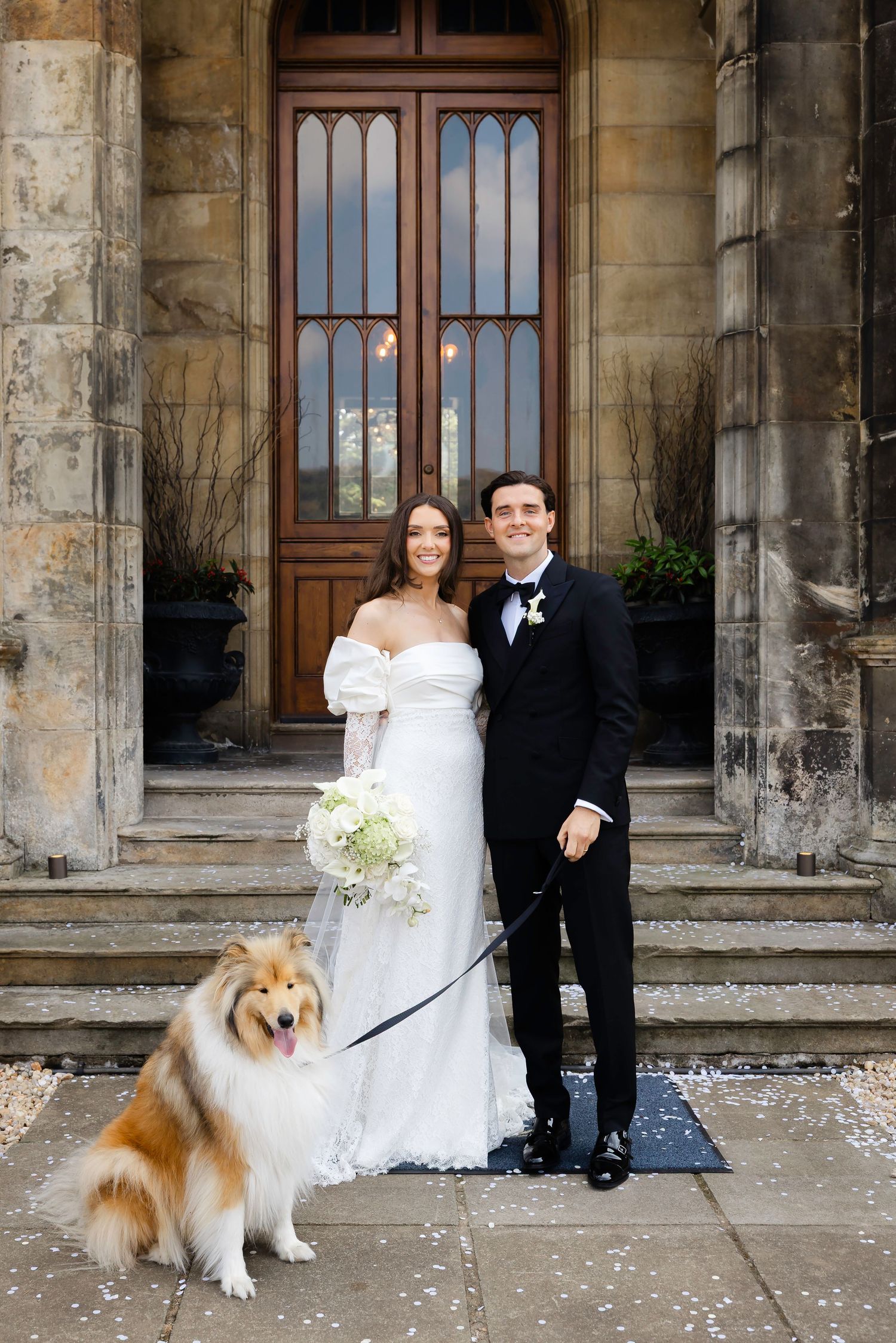 Bride and groom pose with a Collie dog on steps of a stone manor house, bride holding white bouquet.