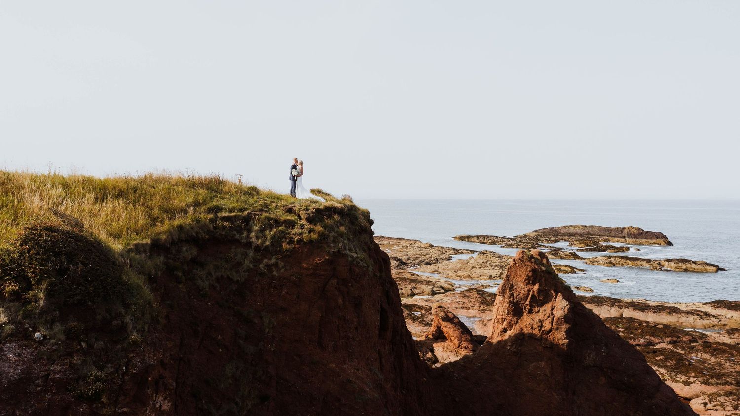 Someone stands on a dramatic coastal cliff edge overlooking the rugged rocky shoreline and ocean at sunset.