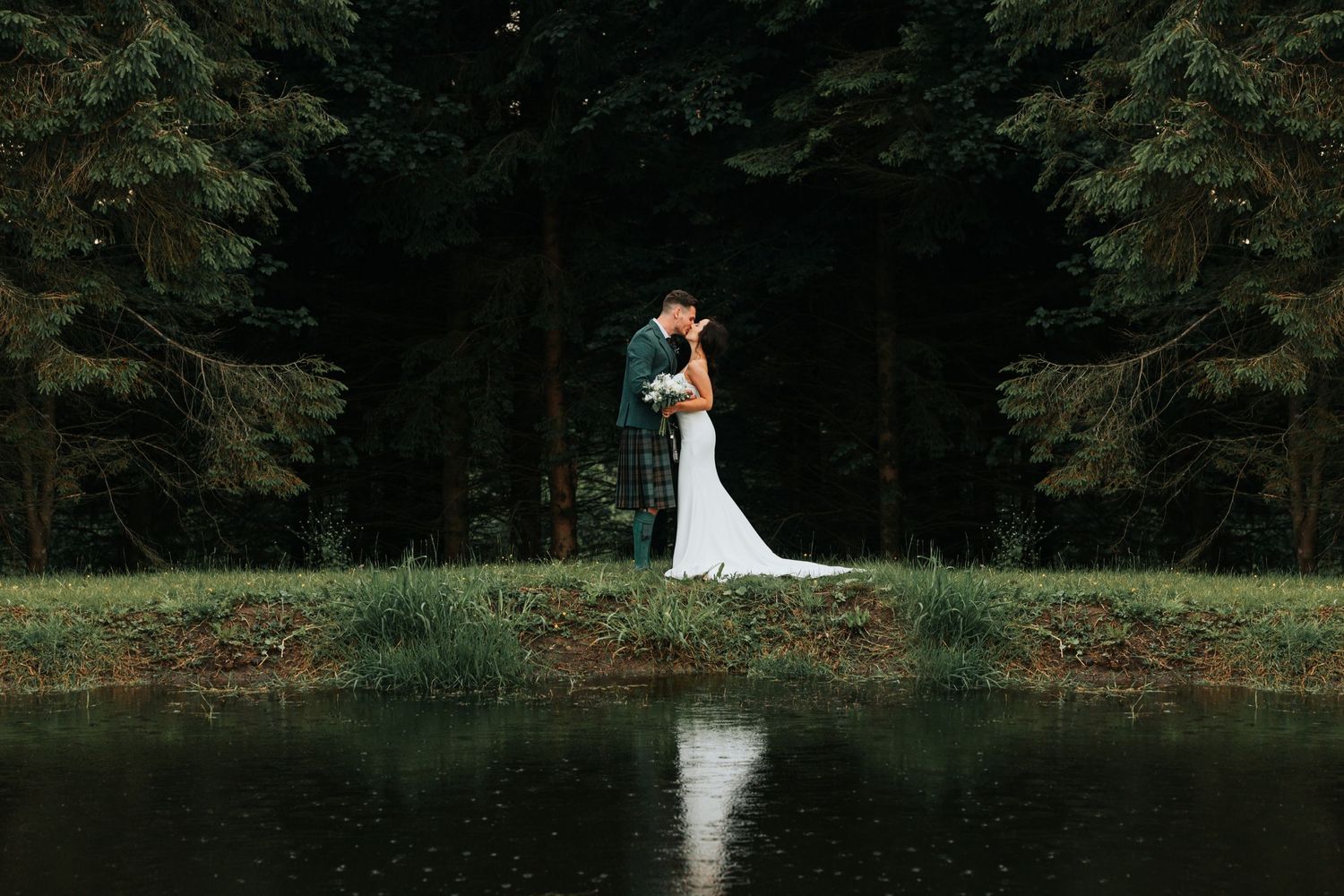 Two figures embrace by a tranquil lake in a forest setting, their reflection mirrored in the still water below.